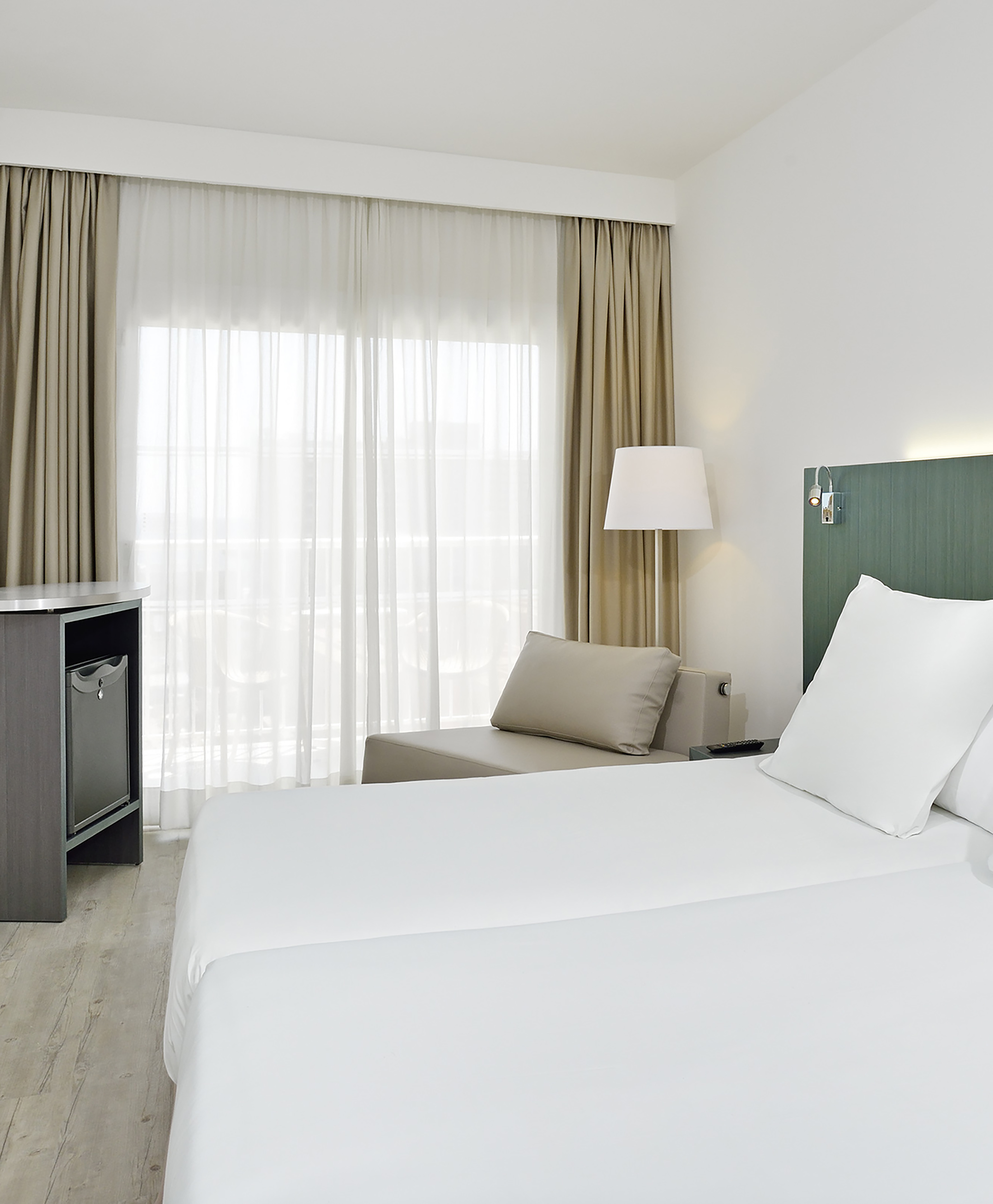 Modern hotel room with white bed, beige lounge, large window, and minibar.