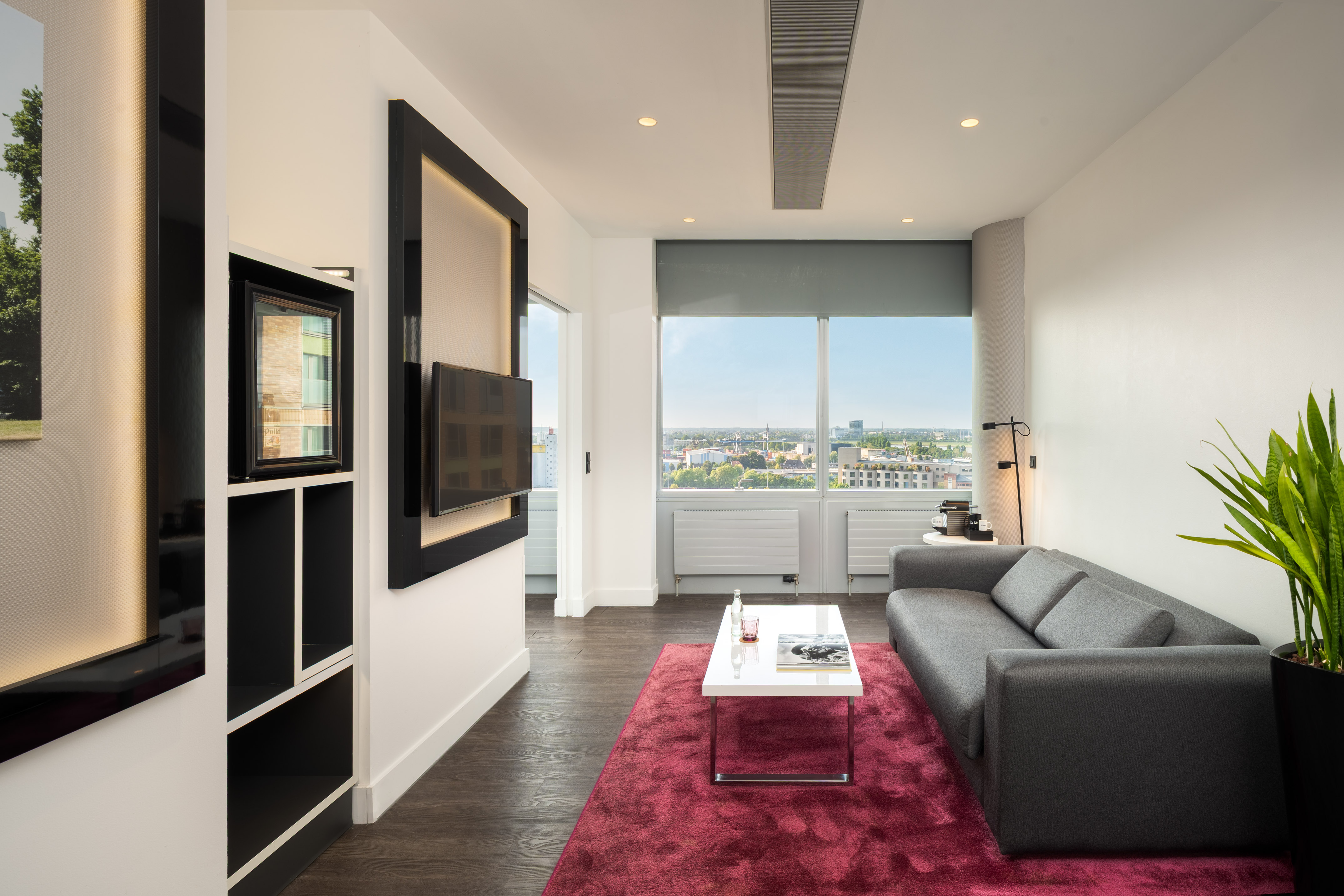 Modern hotel suite living room with city view, gray sofa, red rug, wall TV.