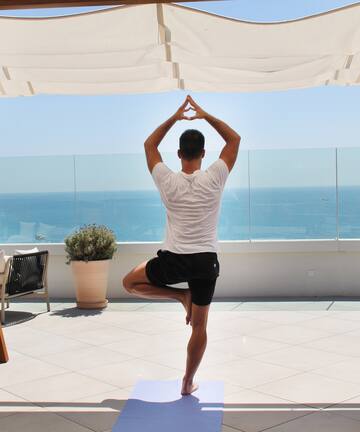 a man standing on one leg on a yoga mat with a body of water in the background