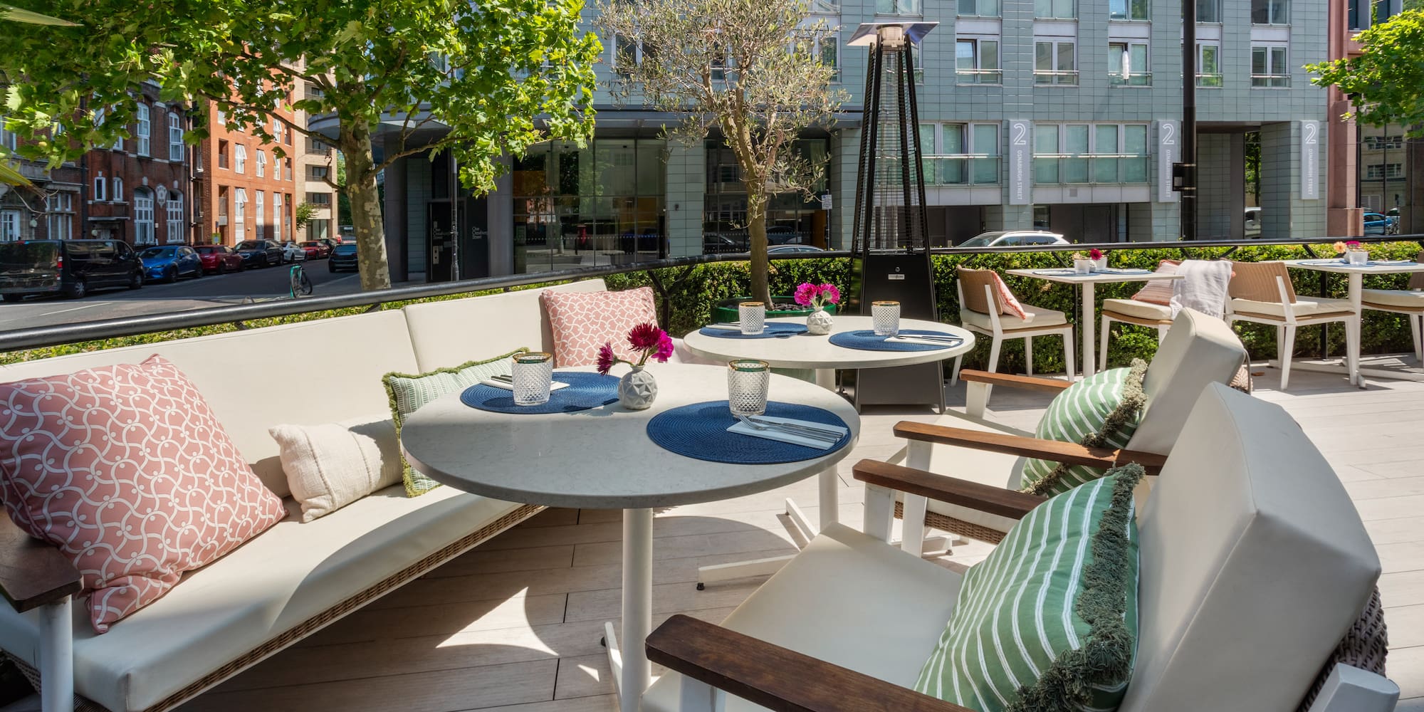 Sunny outdoor dining patio with tables, sofa, greenery, and urban buildings.