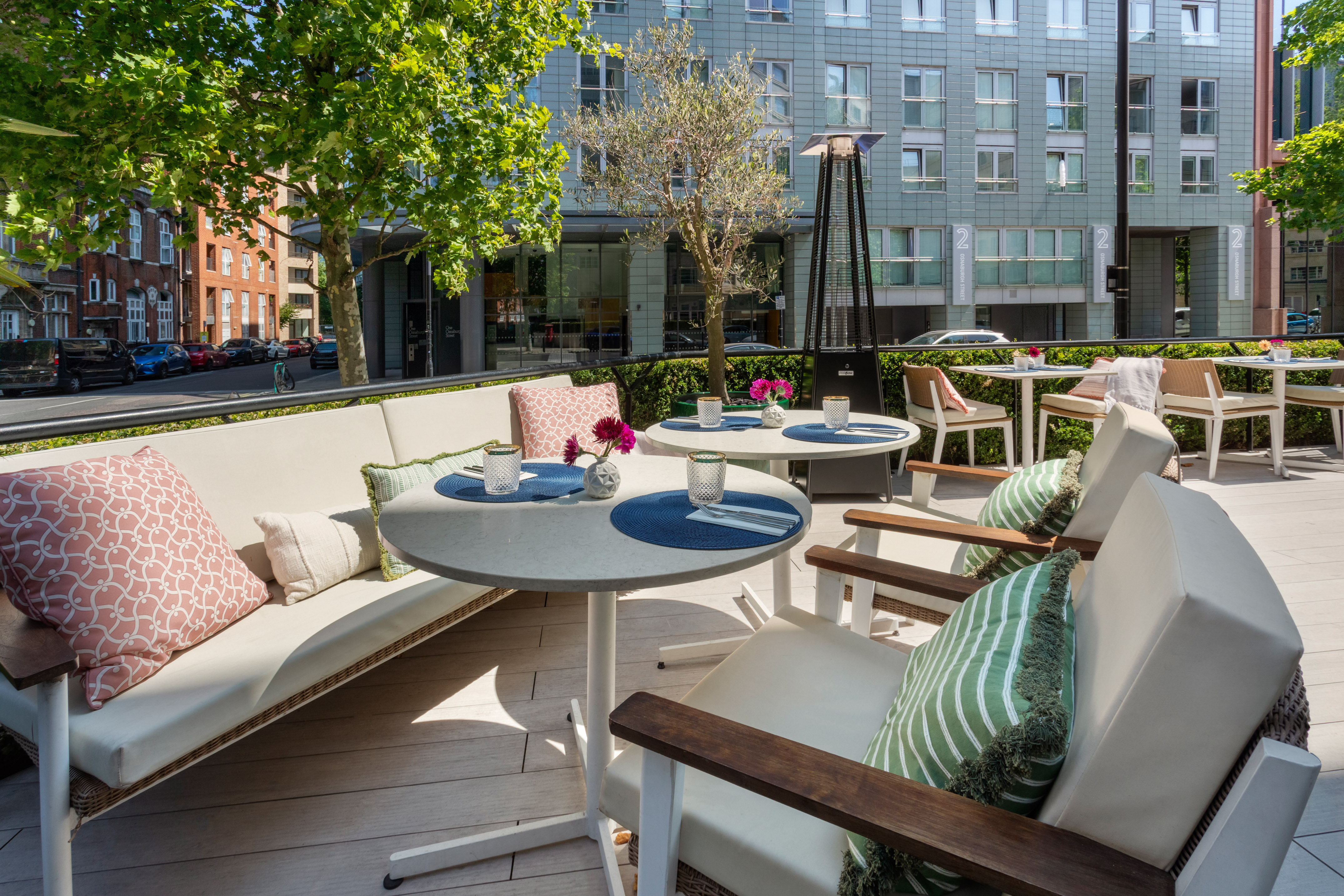 Sunny outdoor dining patio with tables, sofa, greenery, and urban buildings.