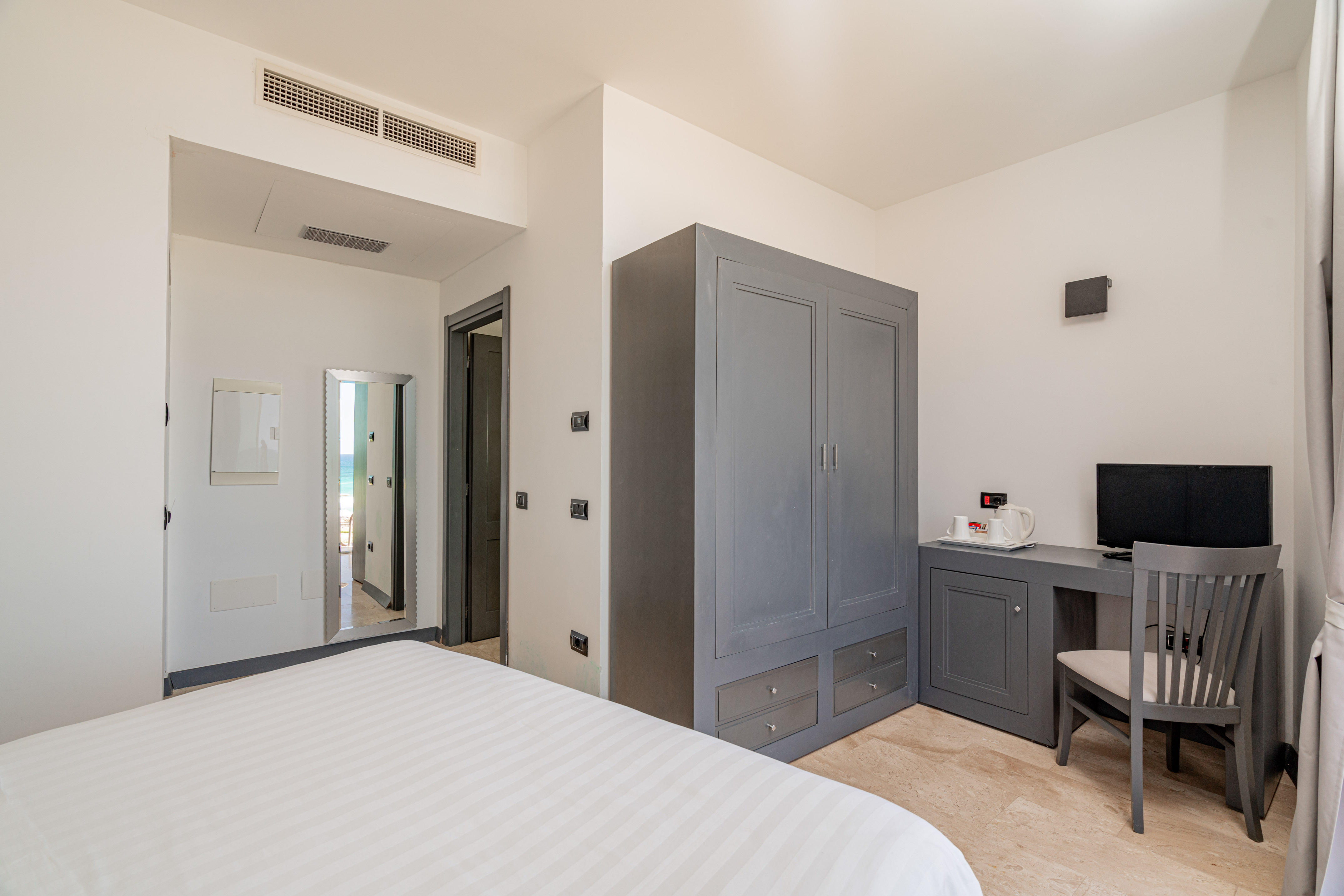Modern hotel bedroom with white bed, gray wardrobe, desk, and ocean view.
