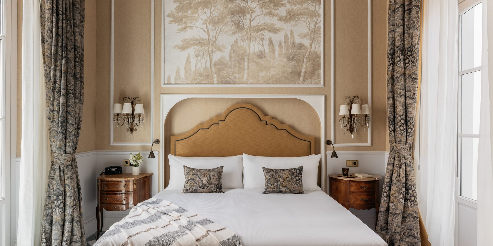Dormitorio elegante, cama con cabecero, mural de árboles y cortinas florales.