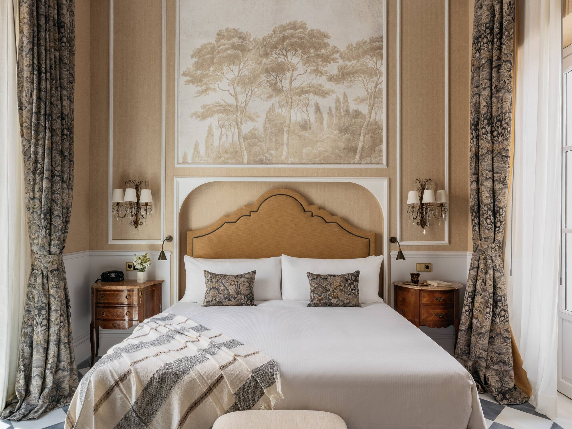 Dormitorio elegante, cama con cabecero, mural de árboles y cortinas florales.