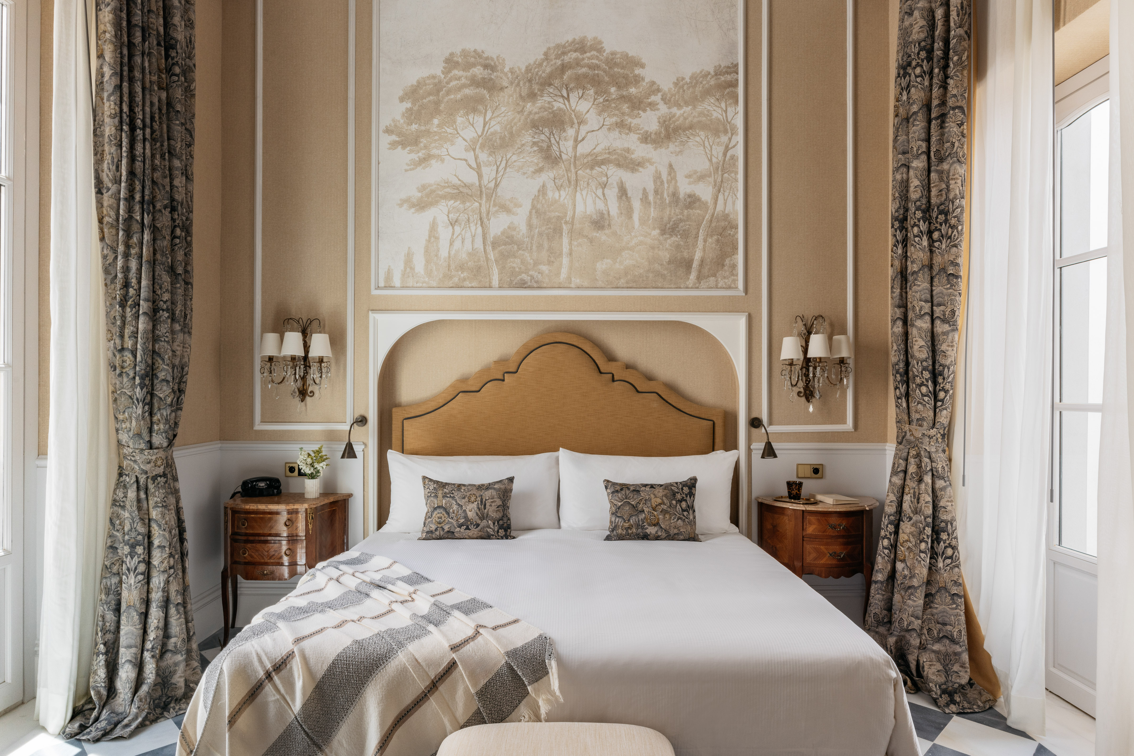Elegant hotel bedroom with large bed, patterned curtains, and scenic tree mural.