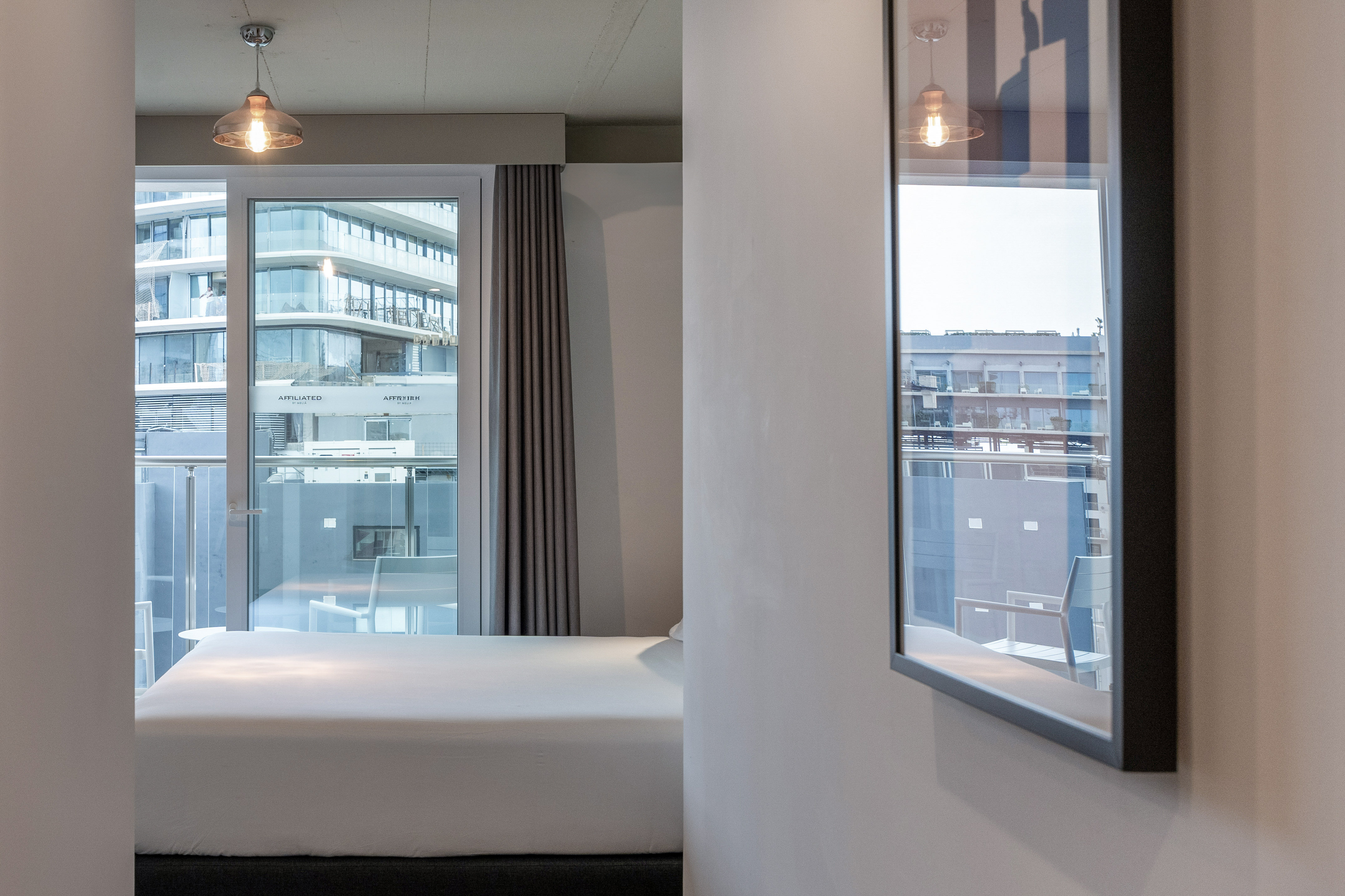 Modern bedroom with white bed, balcony city view, and contemporary lighting.