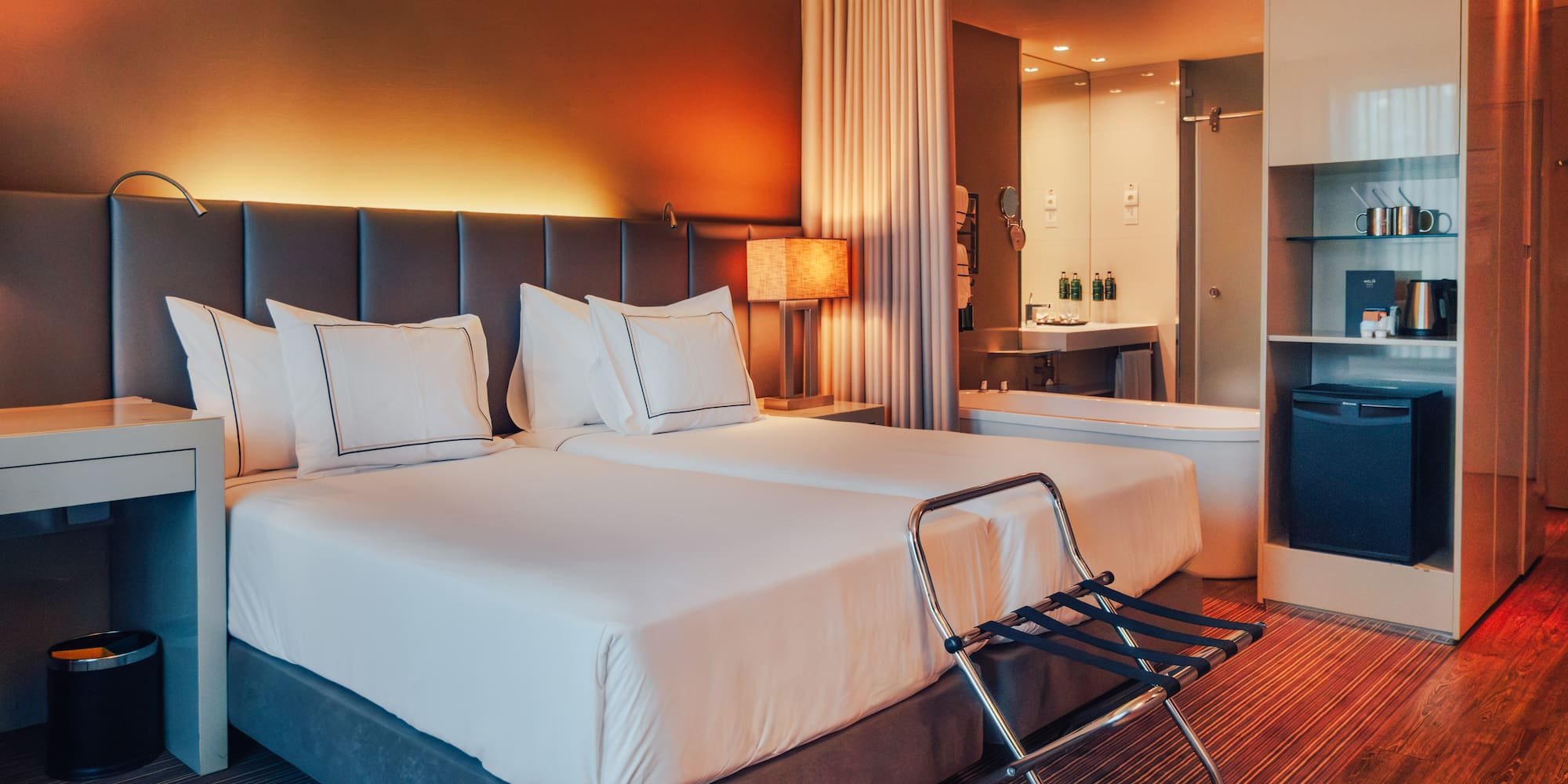 Modern hotel room with twin beds, warm lighting, and open-plan bathroom.