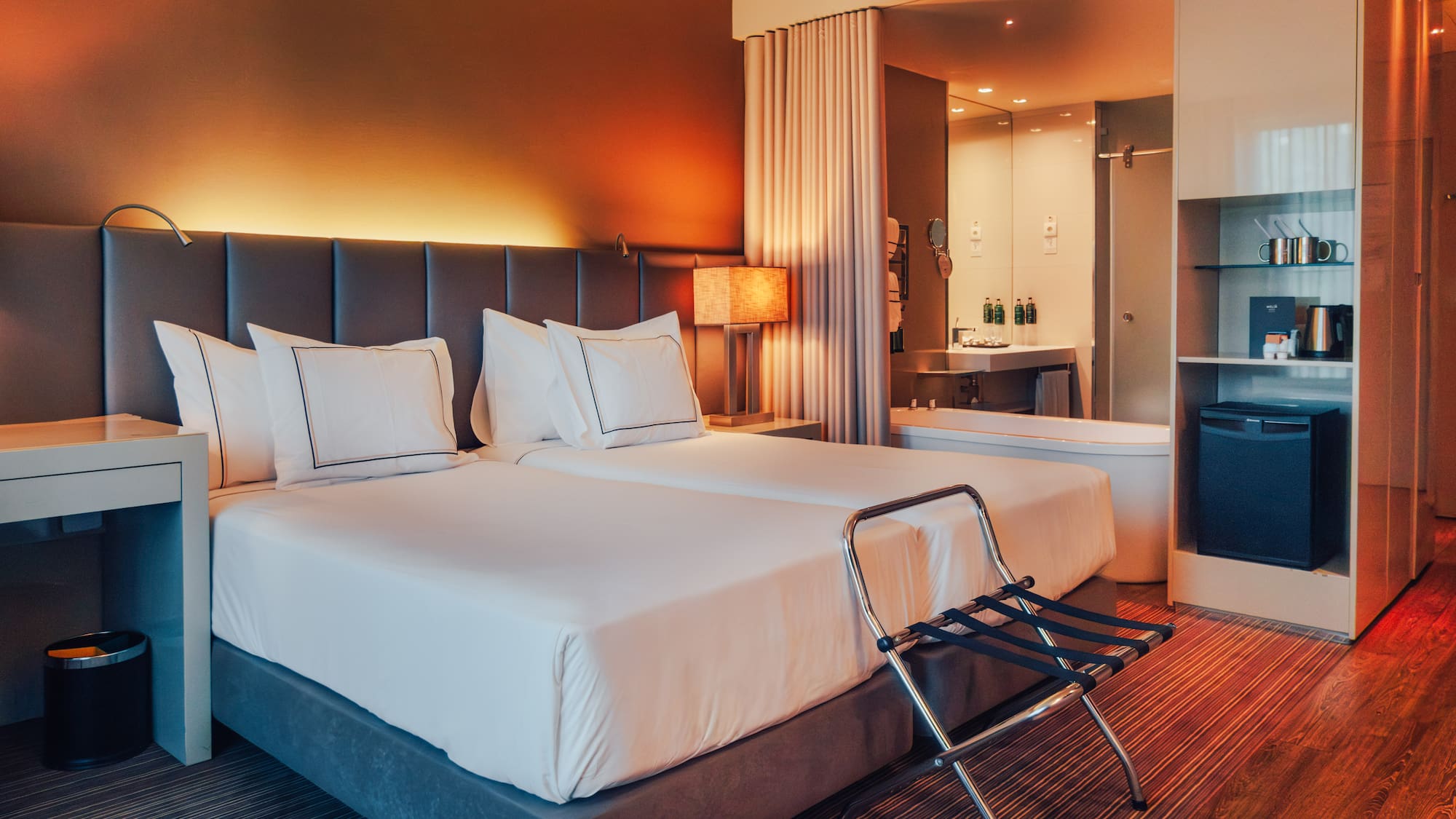 Modern hotel room with twin beds, warm lighting, and open-plan bathroom.