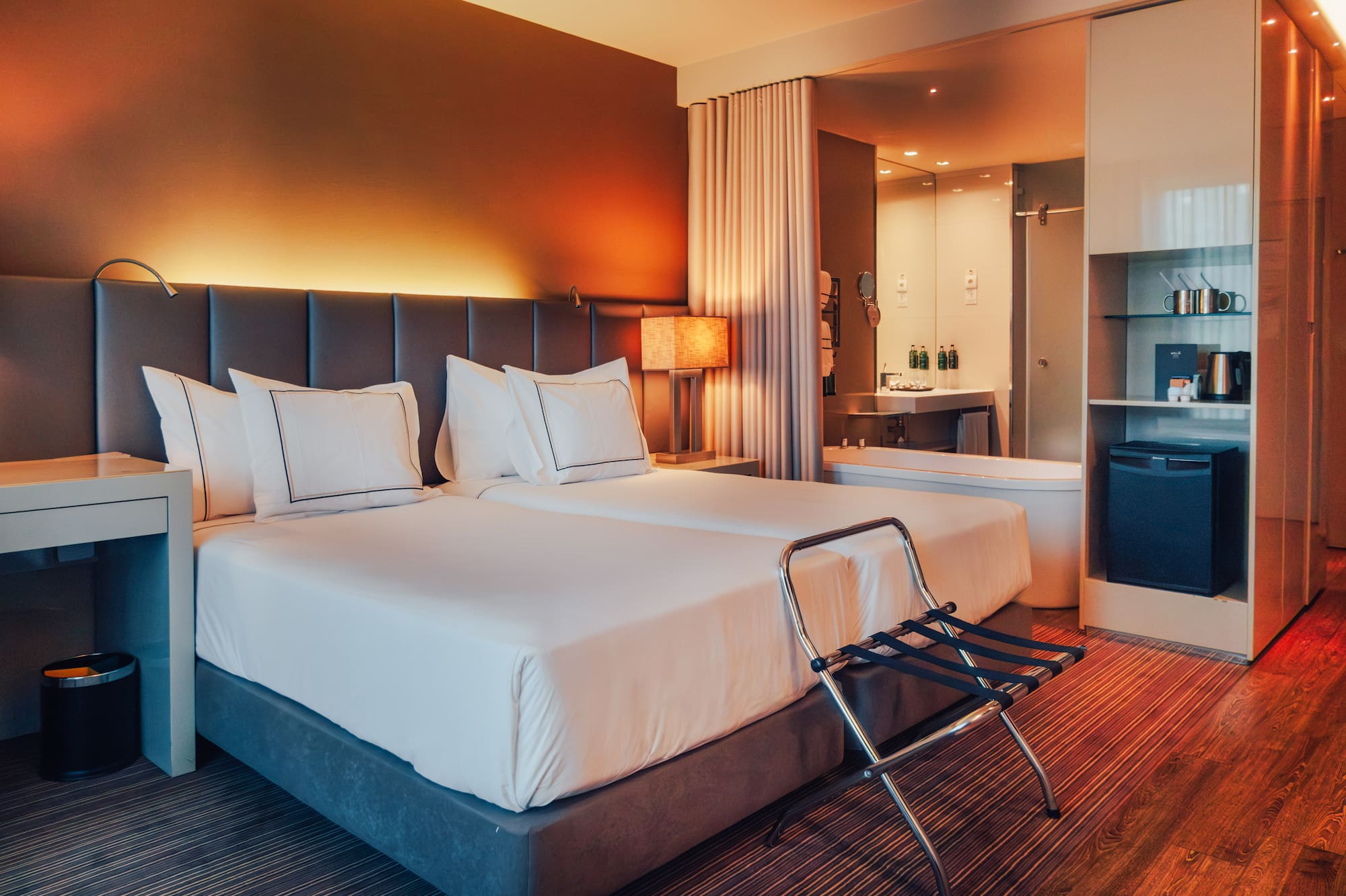 Modern hotel room with twin beds, warm lighting, and open-plan bathroom.