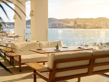 a white couches and tables with a view of the water and mountains in the background