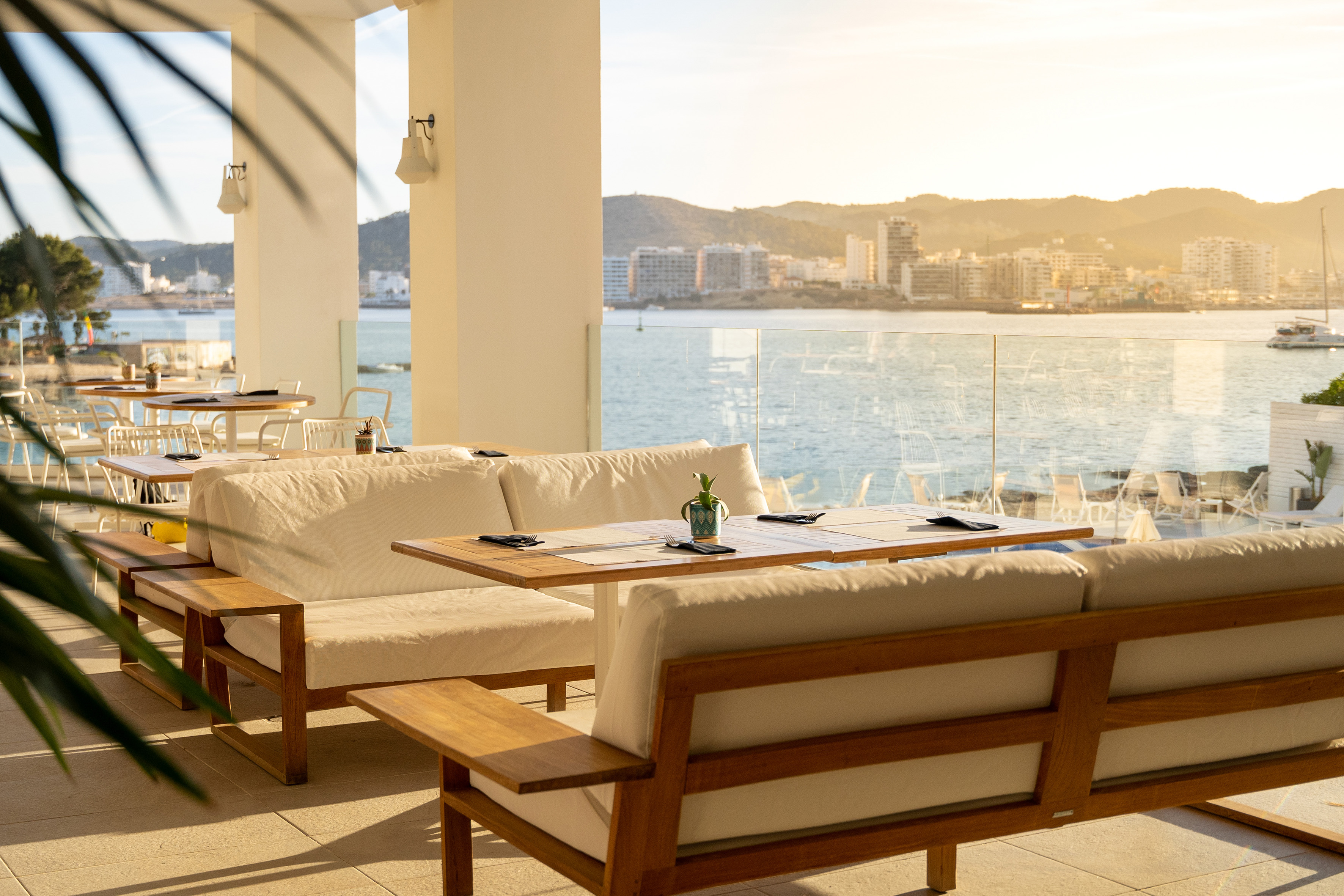 a white couches and tables with a view of the water and mountains in the background