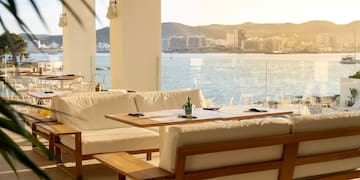a white couches and tables with a view of the water and mountains in the background