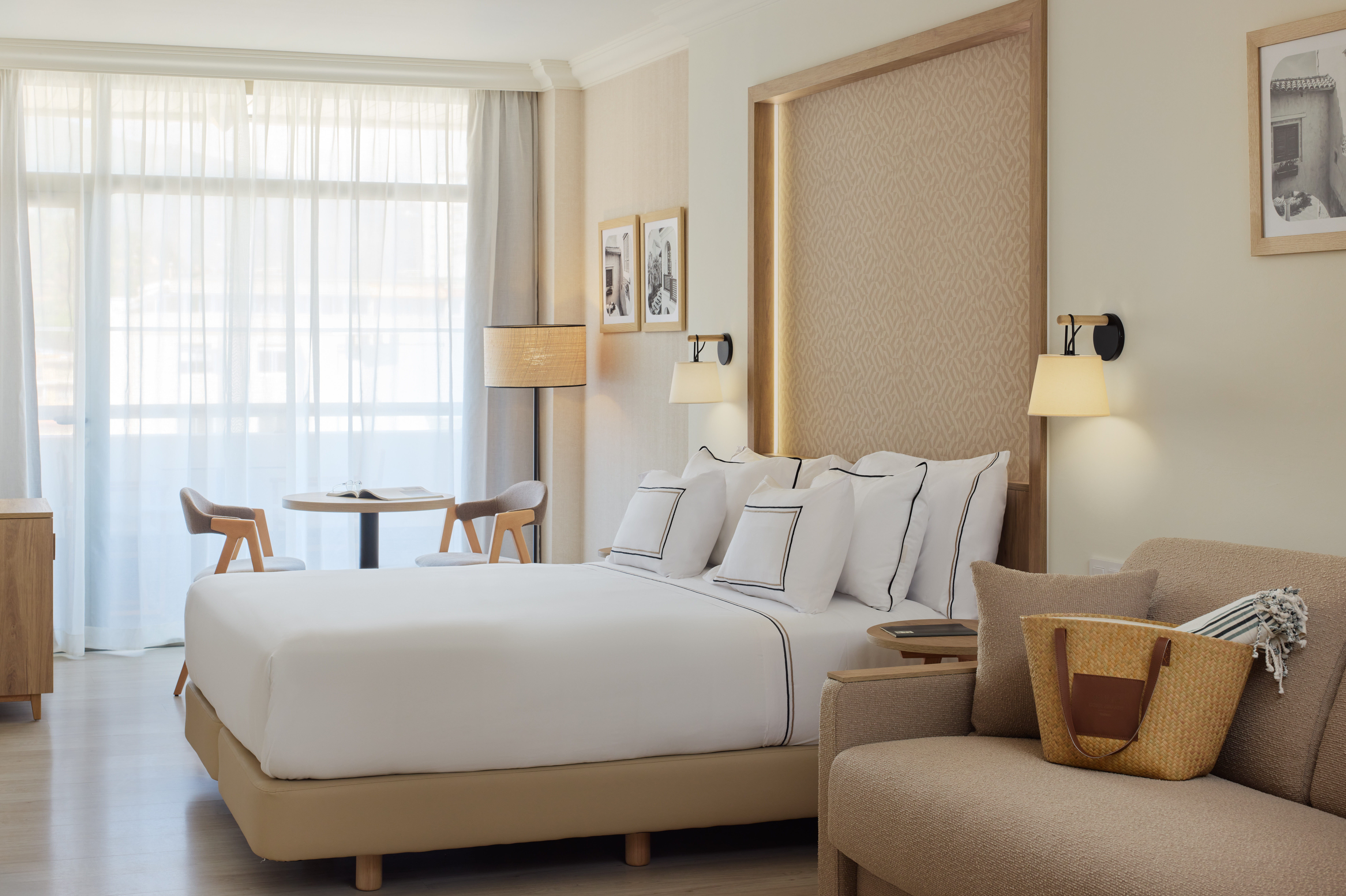 Modern hotel room with bed, sofa, table, and bright window with sheer curtains.