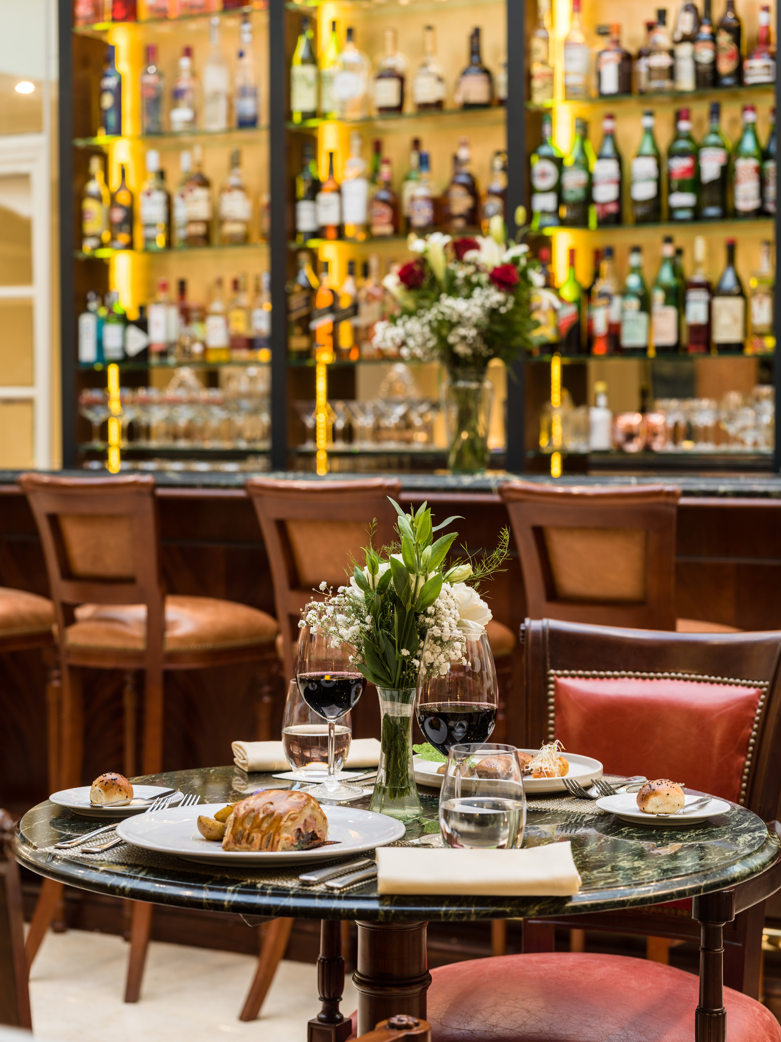 Fine dining table with food, red wine, flowers; elegant restaurant bar in background.
