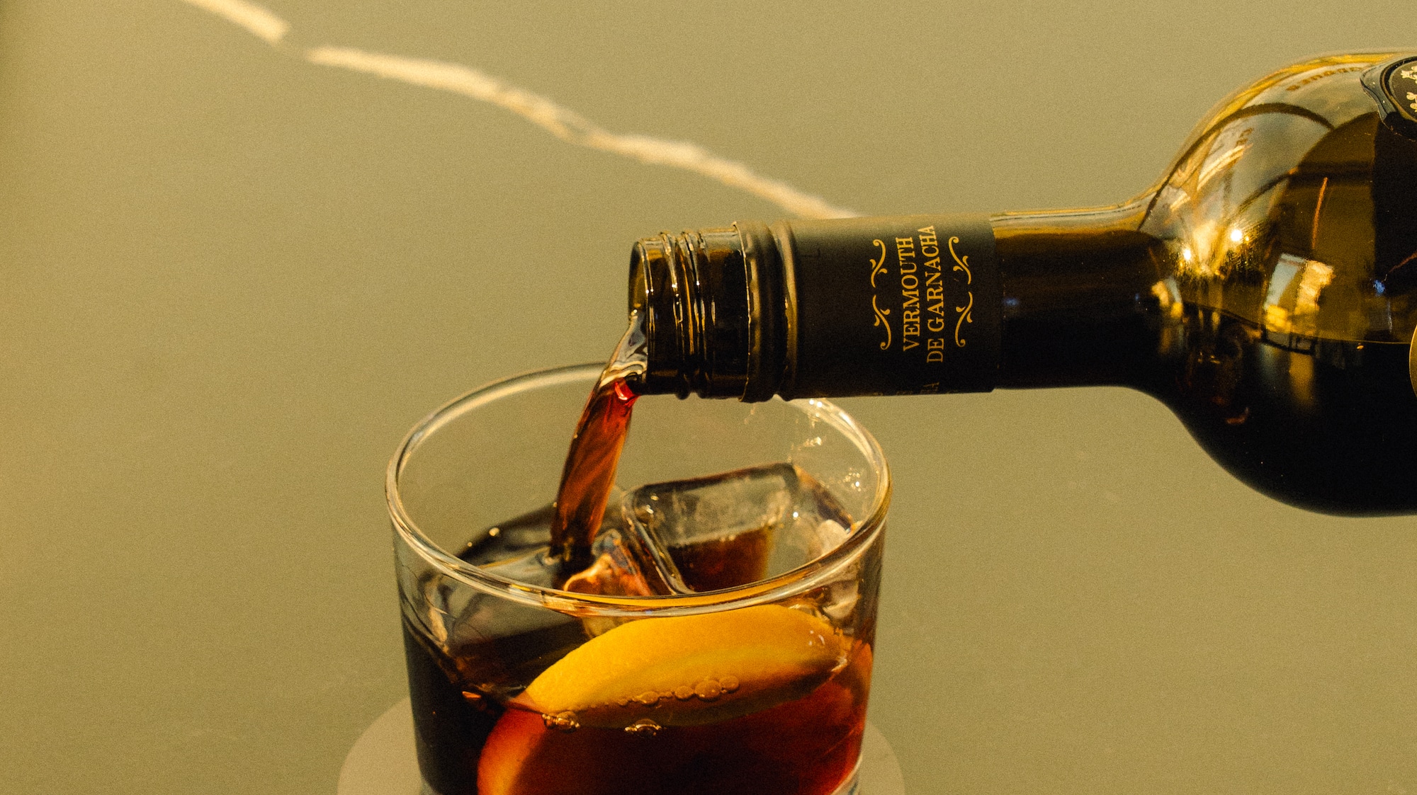 Pouring red vermouth from a Garnacha bottle into a glass with ice and orange.