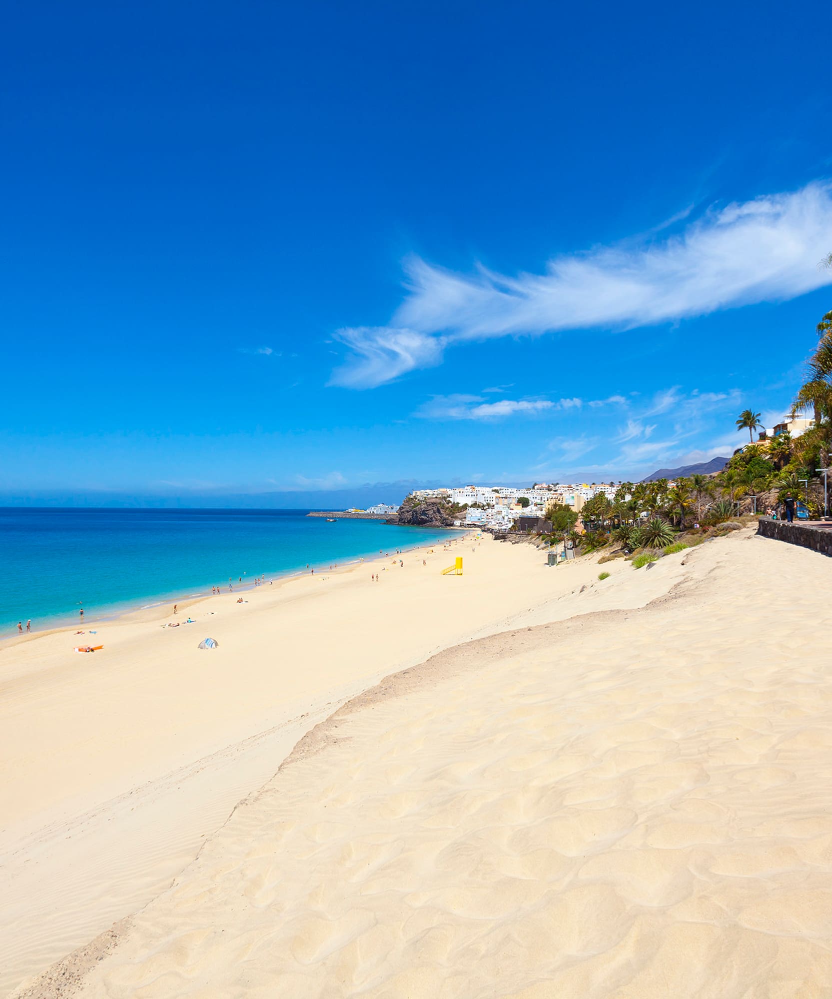 Sunny white sand beach with turquoise ocean, distant town, and palm trees.