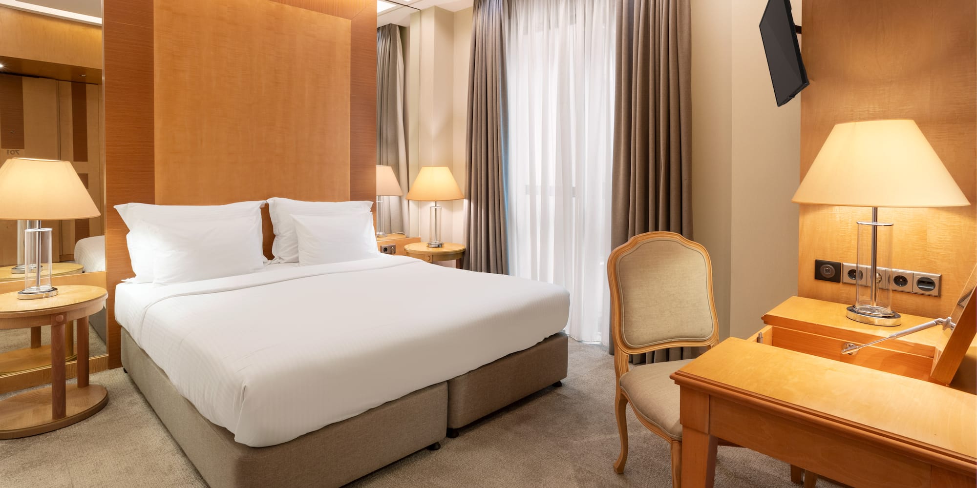 Modern hotel room with king bed, wooden headboard, bedside lamps, desk, and TV.