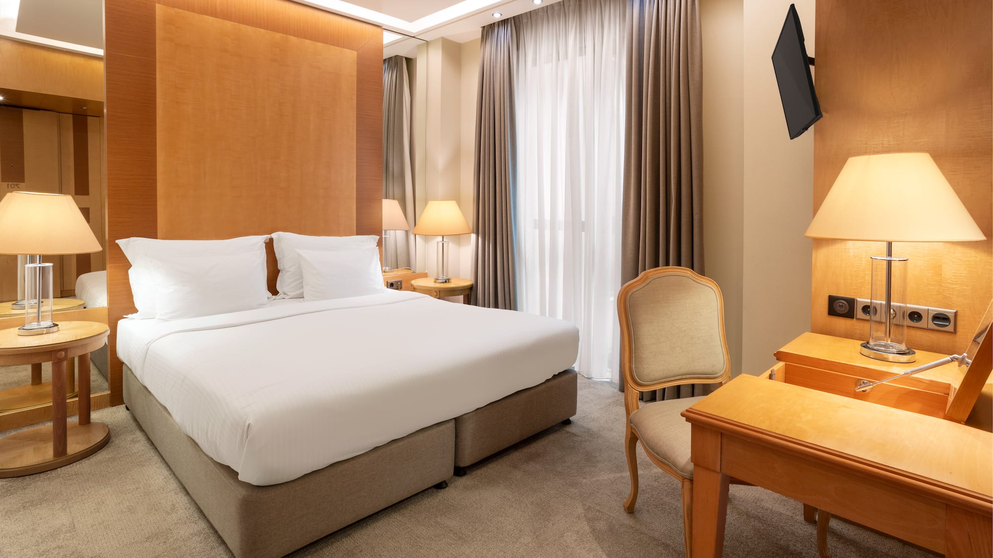 Modern hotel room with king bed, wooden headboard, bedside lamps, desk, and TV.