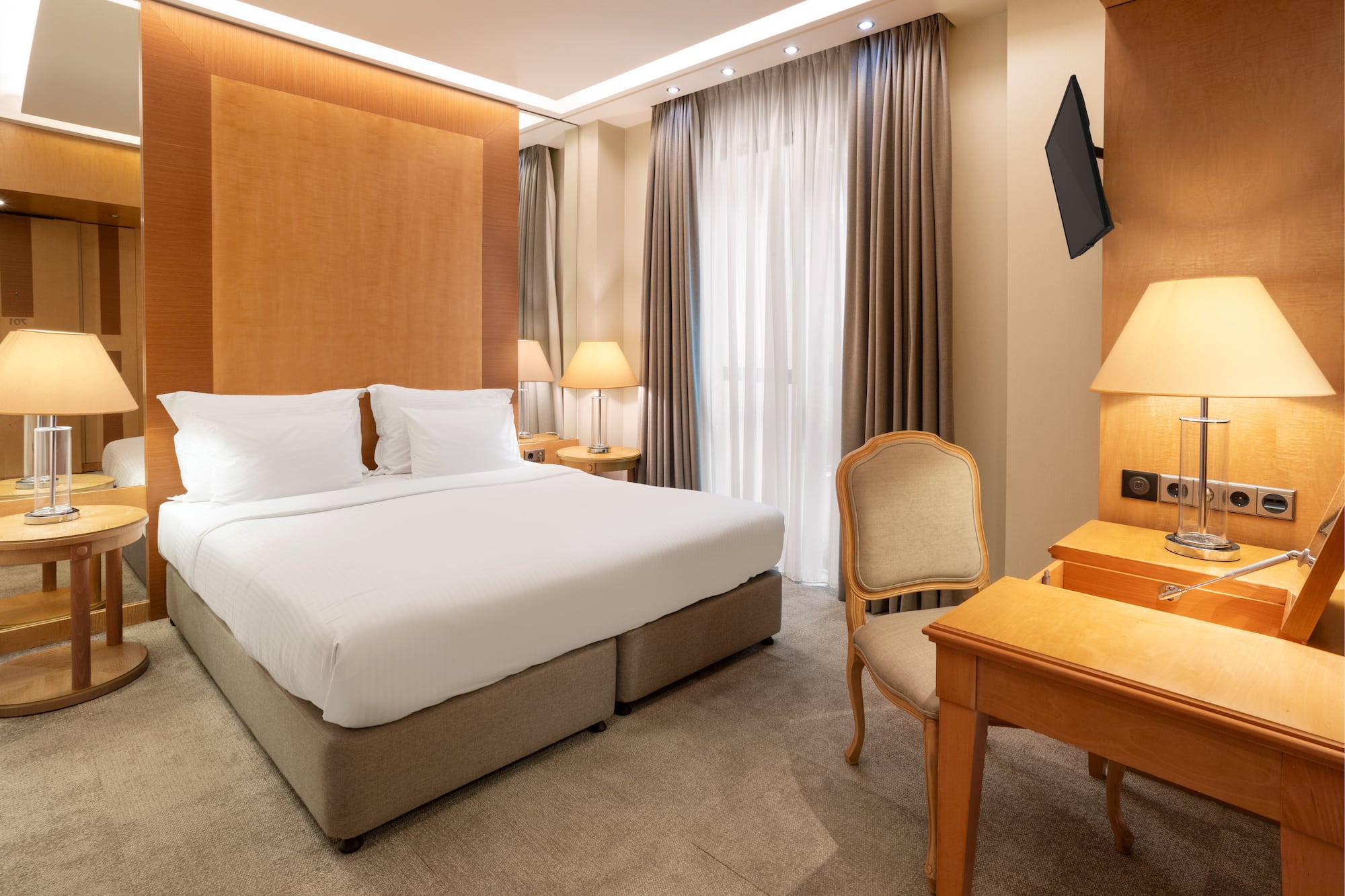 Modern hotel room with king bed, wooden headboard, bedside lamps, desk, and TV.