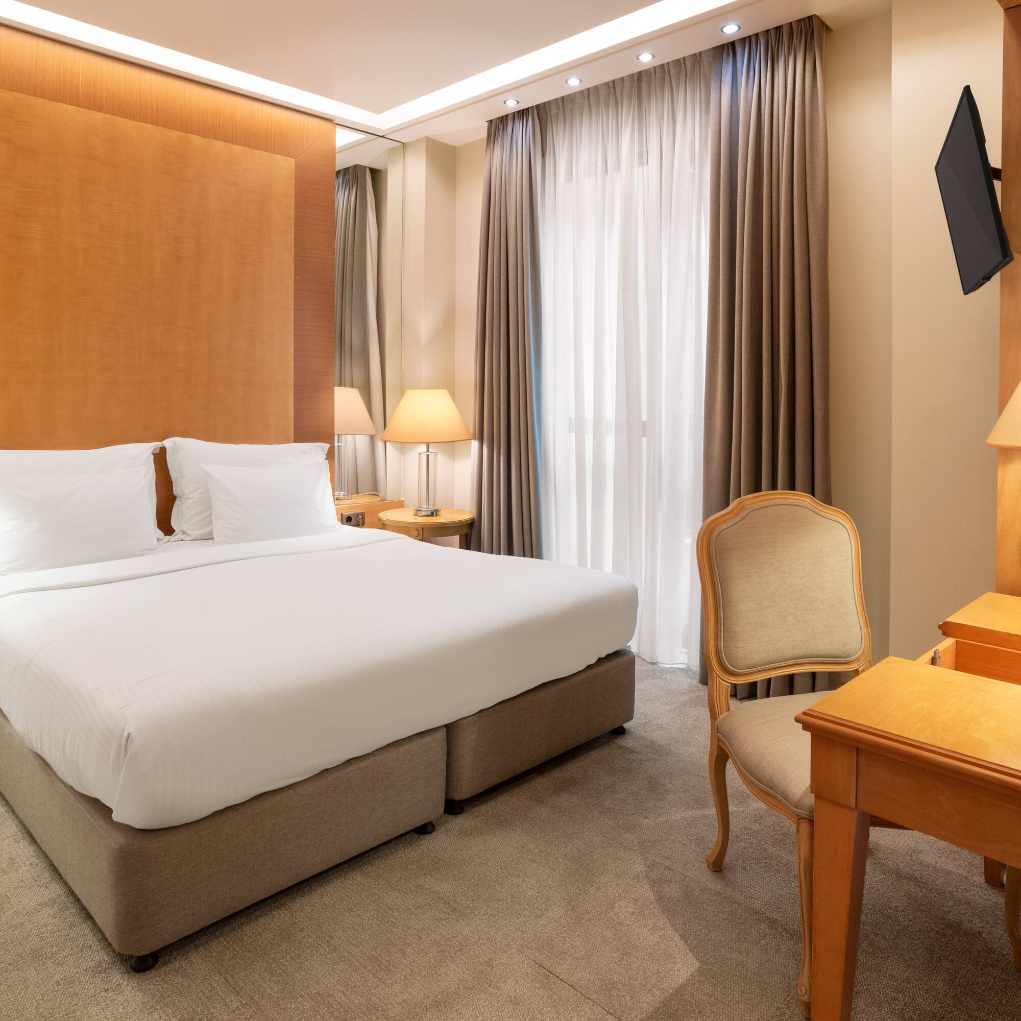 Modern hotel room with king bed, wooden headboard, bedside lamps, desk, and TV.