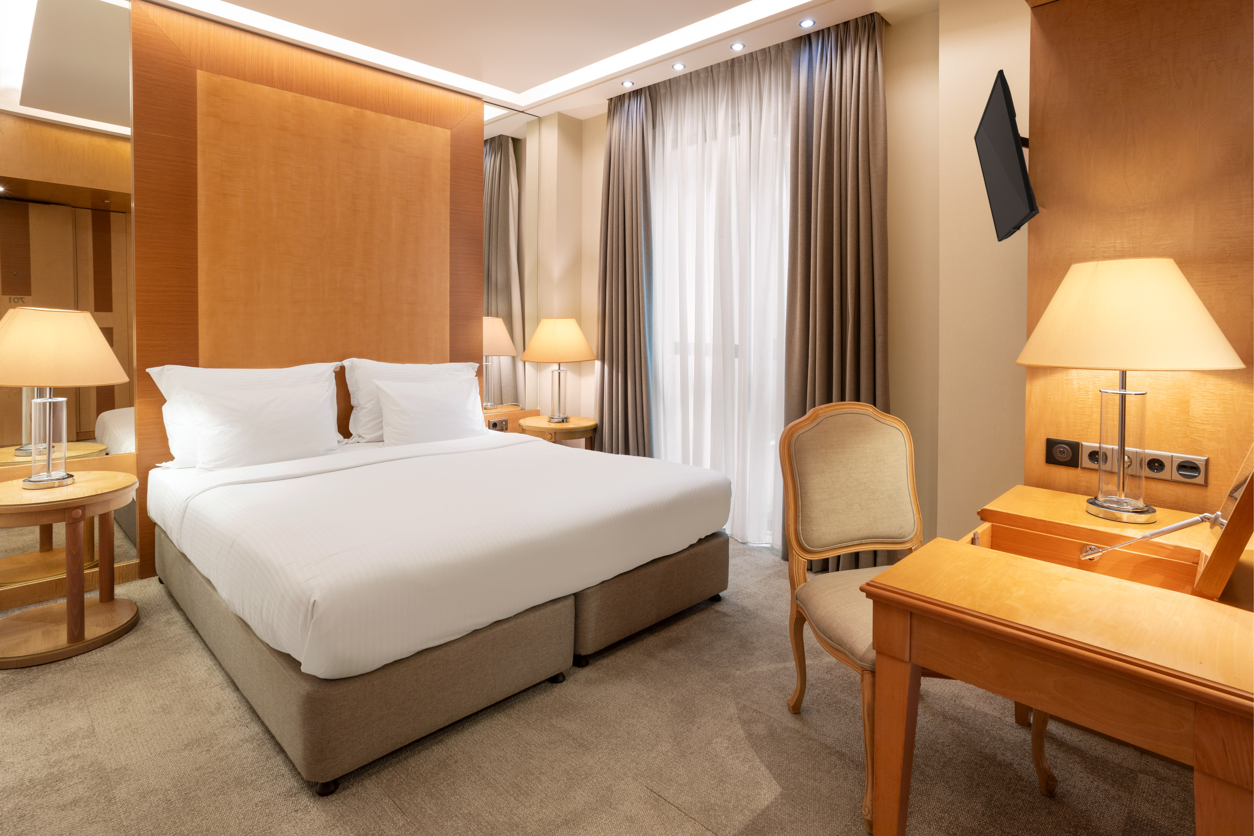 Modern hotel room with king bed, wooden headboard, bedside lamps, desk, and TV.