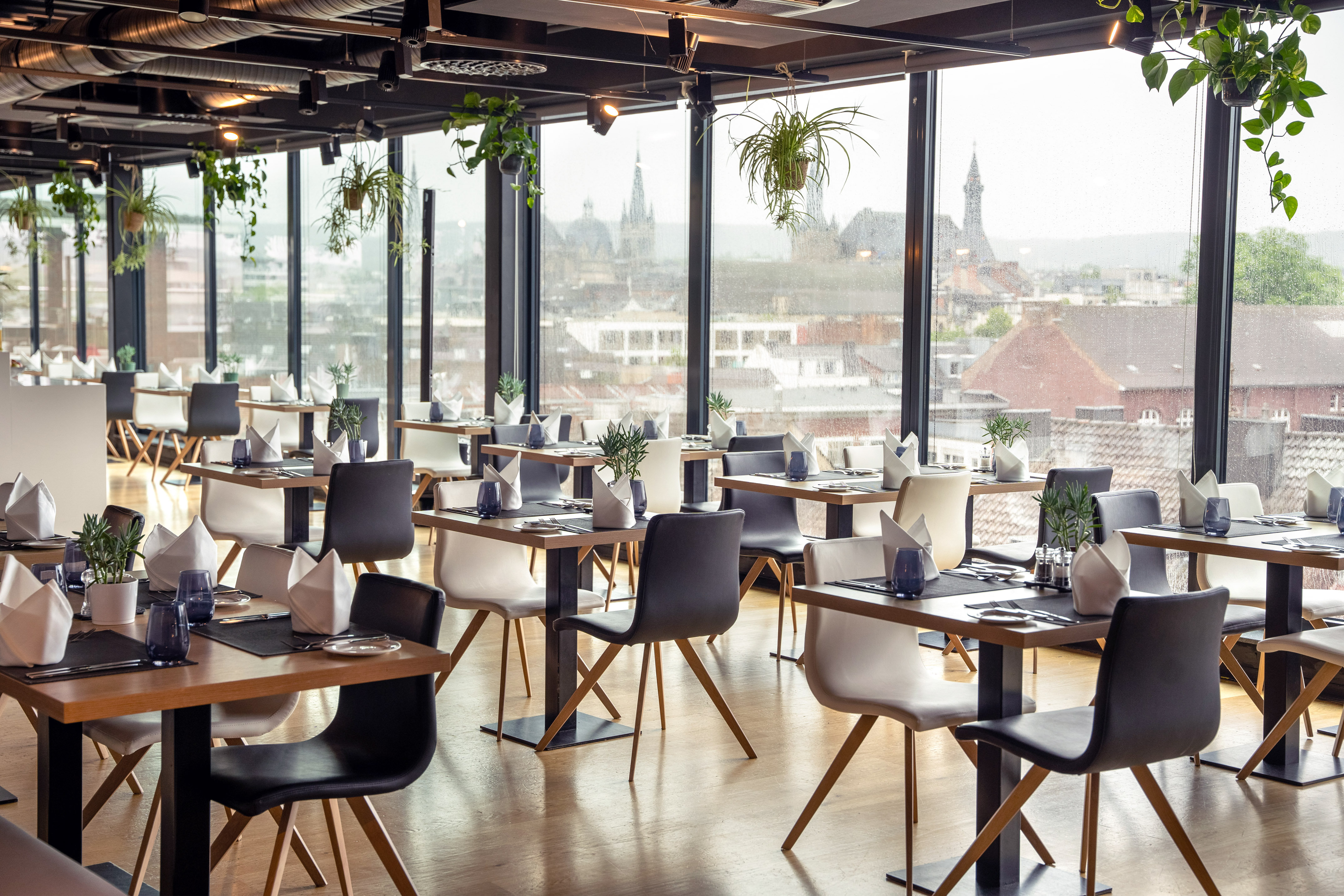 Modern restaurant dining room with city view, elegant tables, and hanging plants.