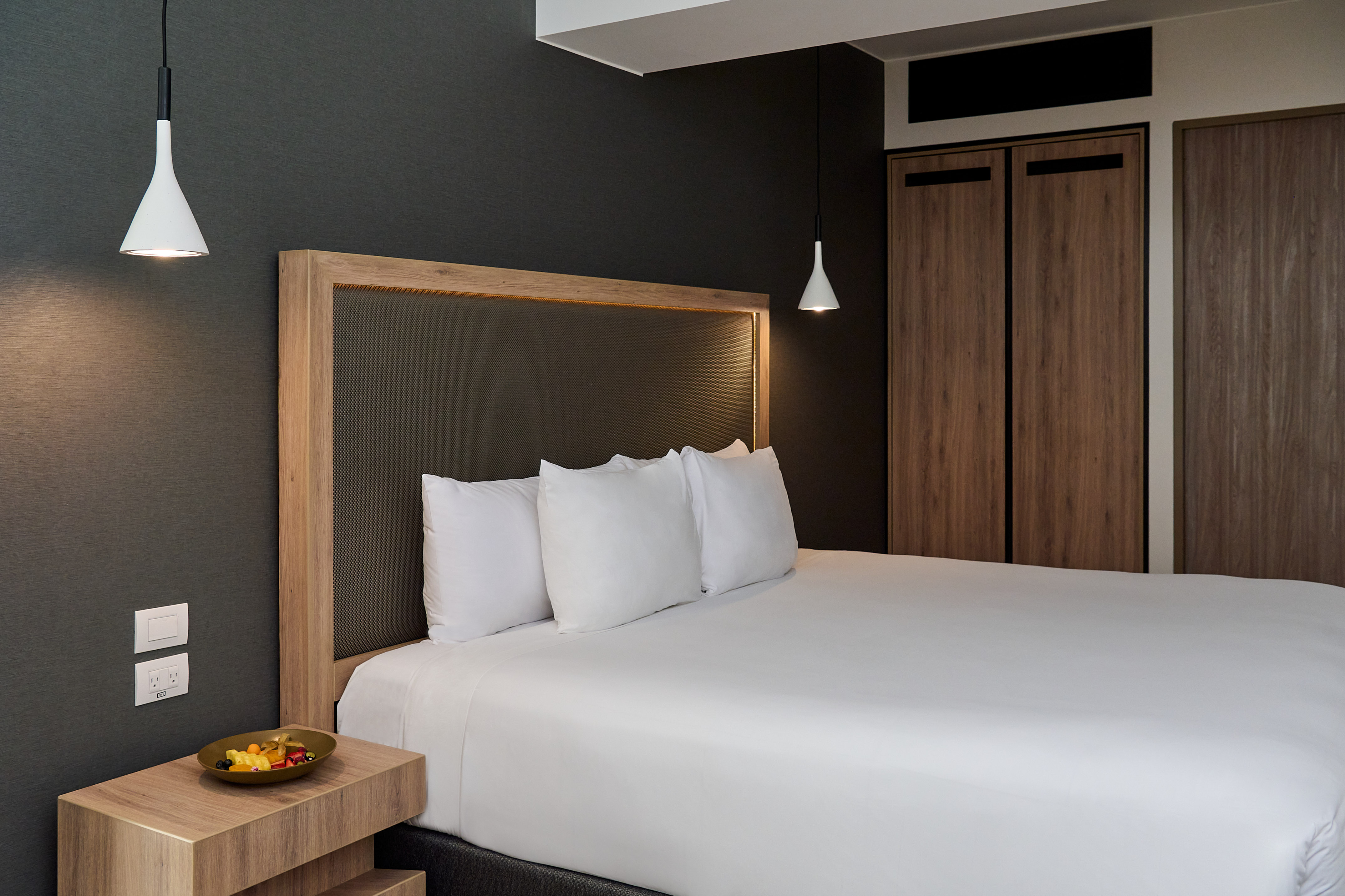 Modern hotel bedroom with dark walls, wood headboard, and white bed.