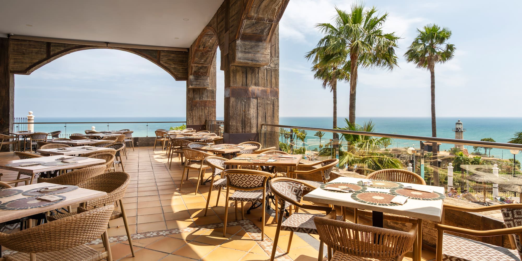 Coastal restaurant terrace with ocean view, palm trees, and a lighthouse.