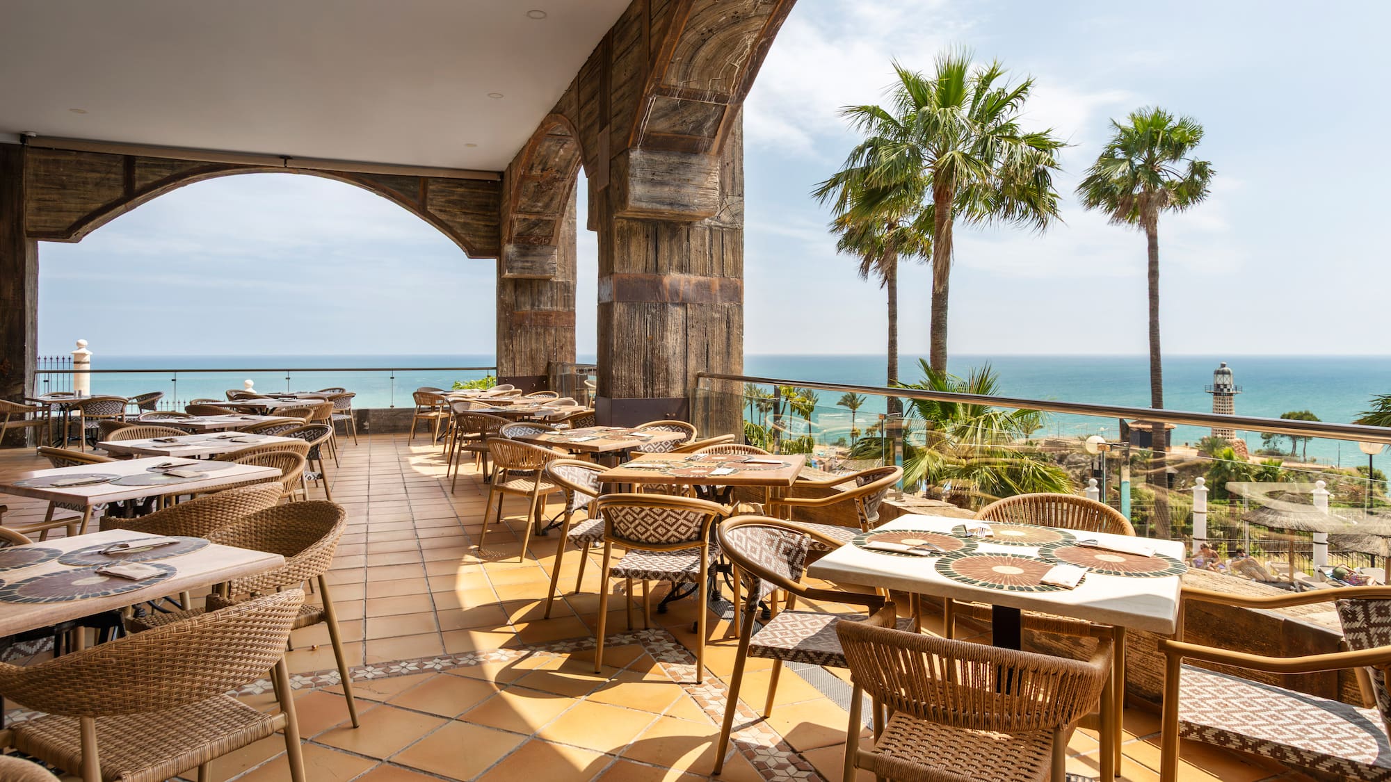 Oceanfront restaurant patio with wooden arches, palm trees, and a distant lighthouse.