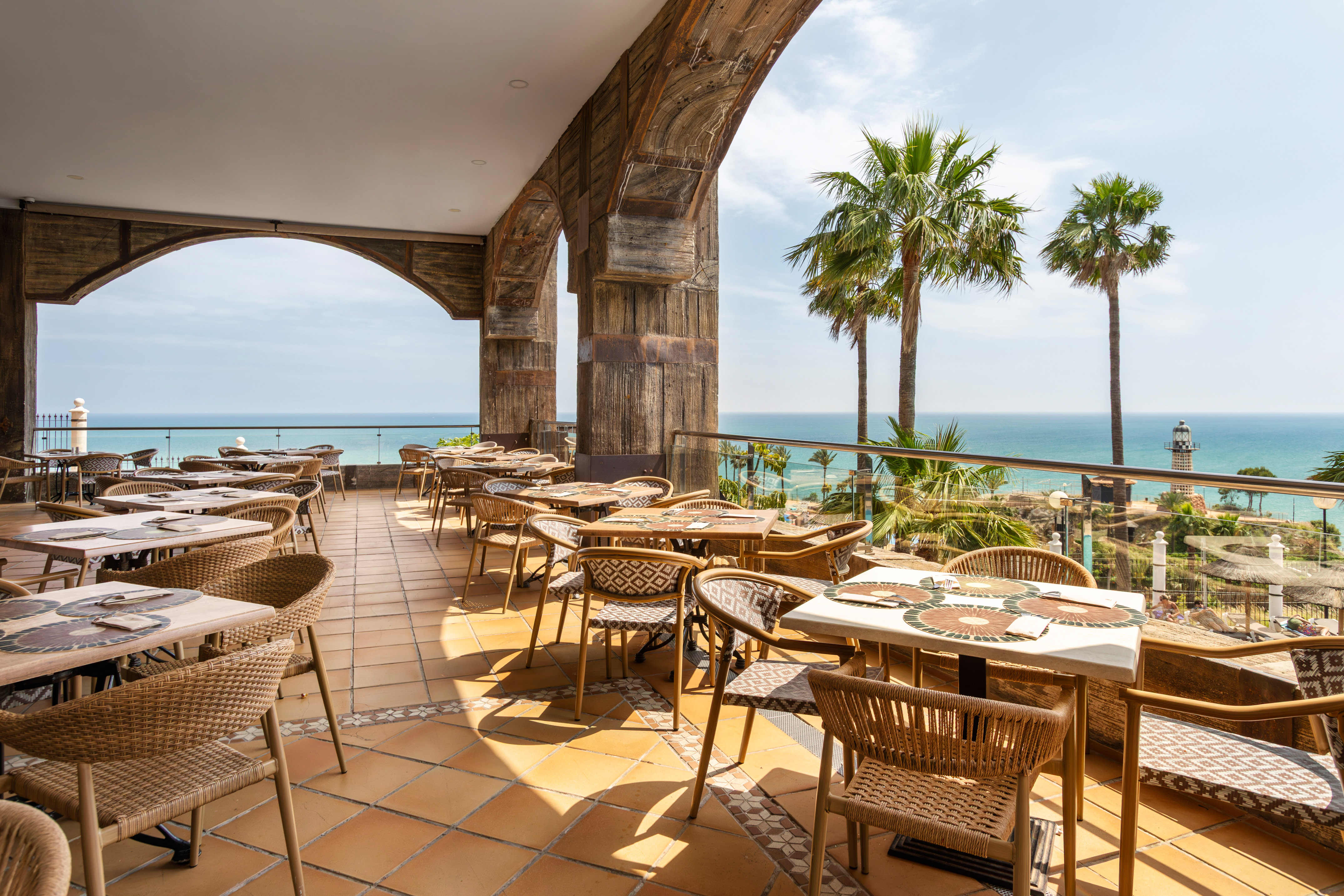 Coastal restaurant terrace with ocean view, palm trees, and a lighthouse.