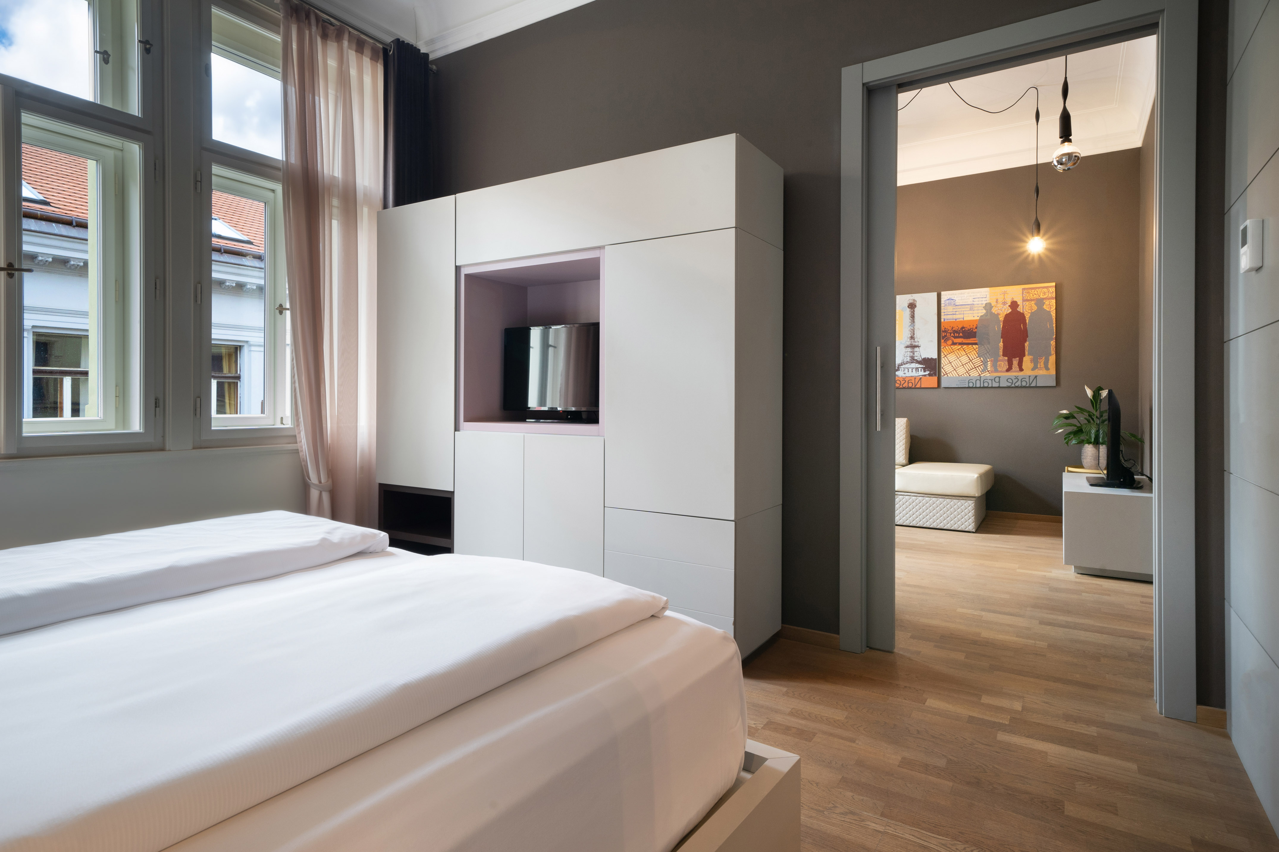 Bright bedroom with white bed, built-in TV wardrobe, and large windows.
