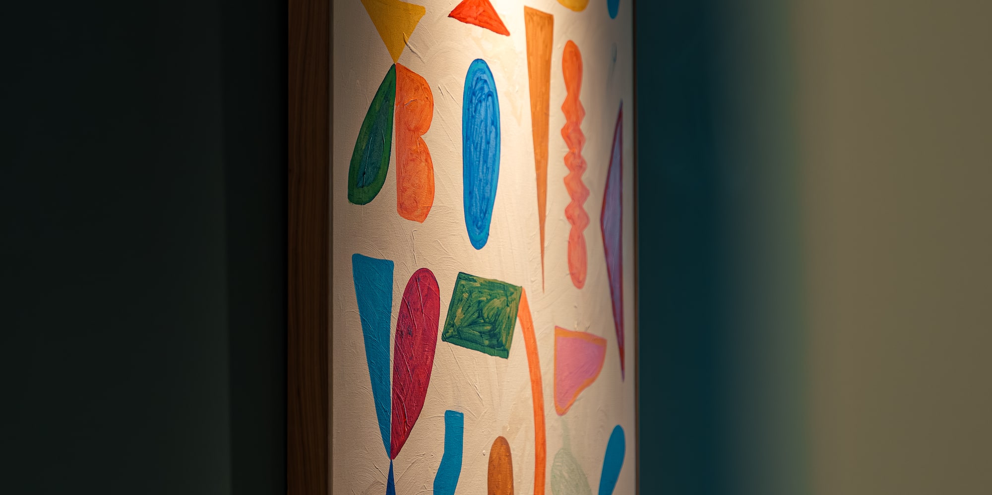 Abstract painting with colorful organic shapes, illuminated on a dark teal wall.