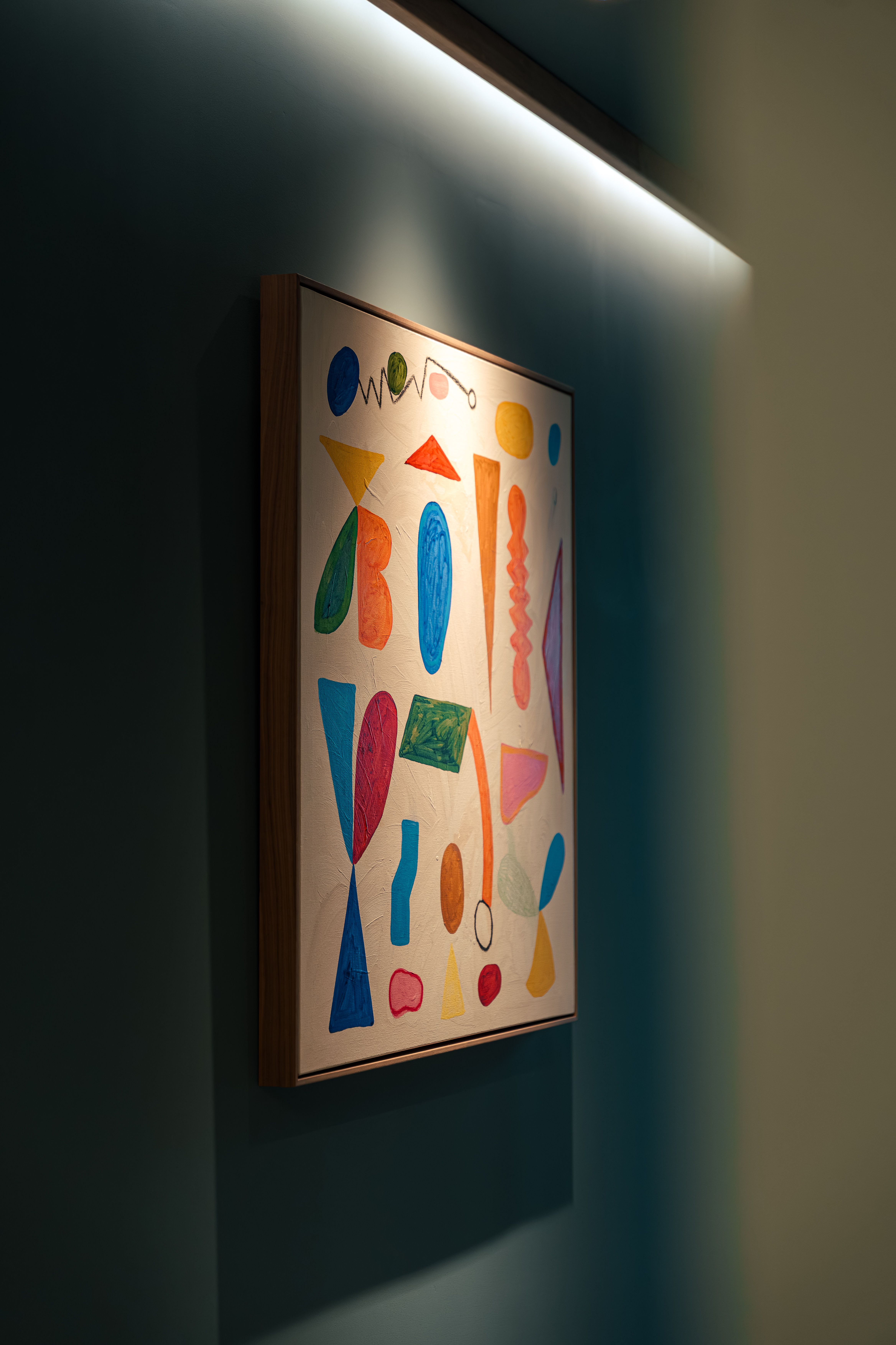 Abstract painting with colorful organic shapes, illuminated on a dark teal wall.