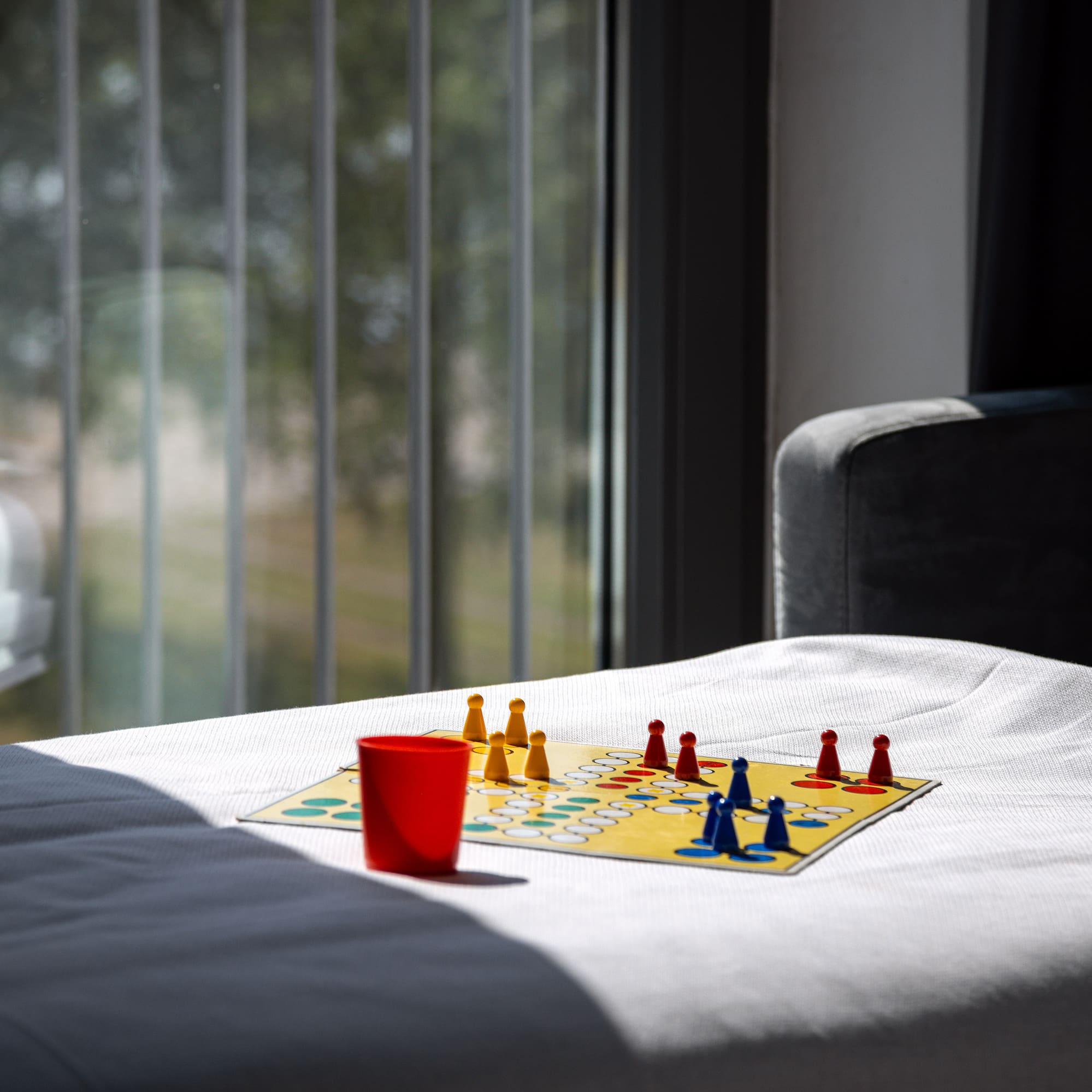 Colorful Ludo board game on white sofa with red cup, window in background.