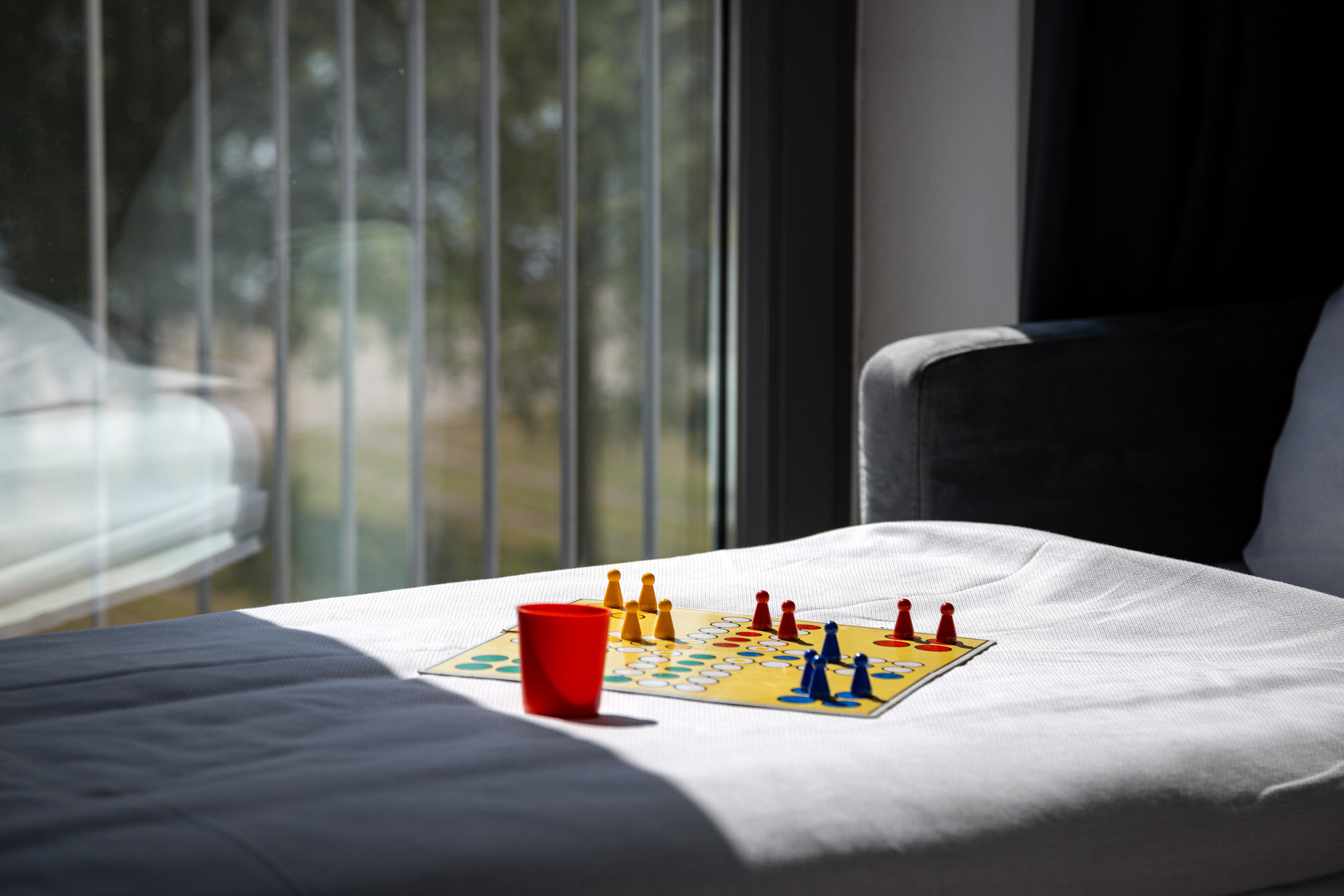 Colorful Ludo board game on white sofa with red cup, window in background.