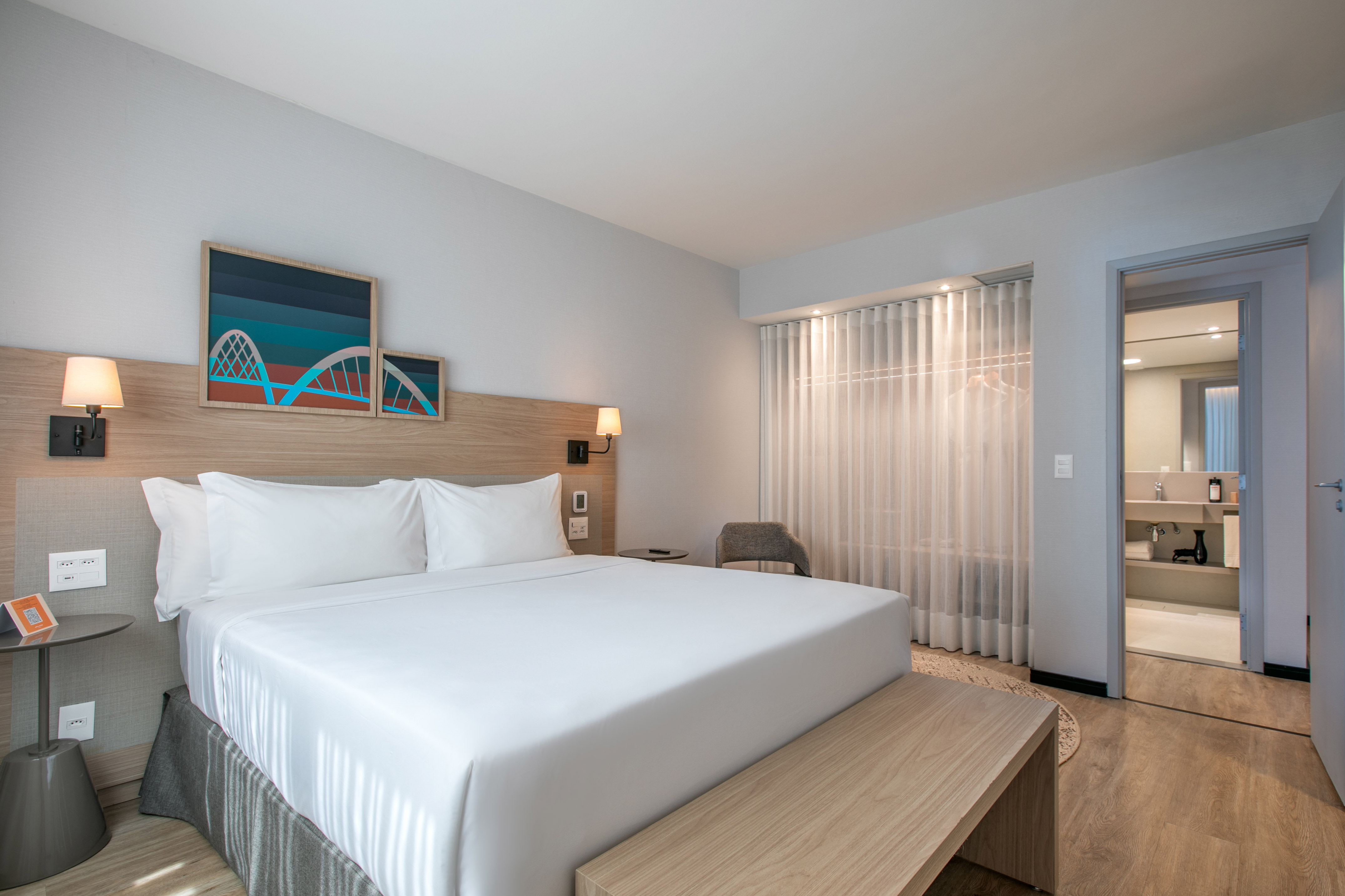 Modern hotel room with king bed, abstract art, wood accents, and bathroom entry.