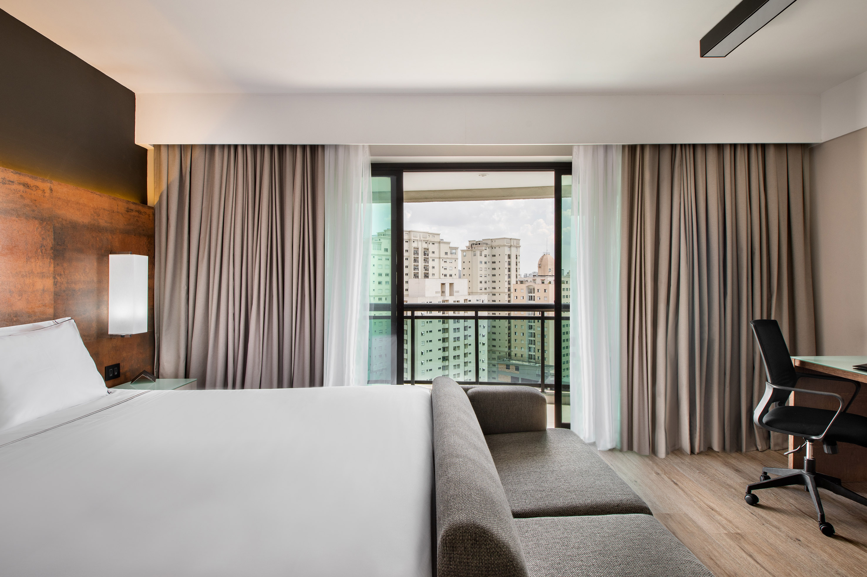 Modern hotel room with king bed, grey sofa, desk, and city view.