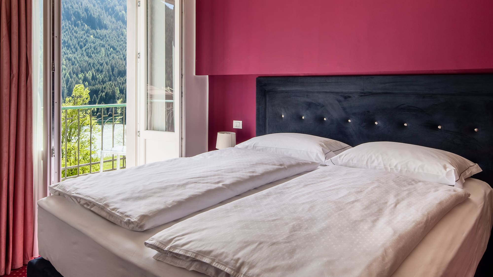Hotel room with magenta wall, bed, and balcony overlooking a forested mountain lake.