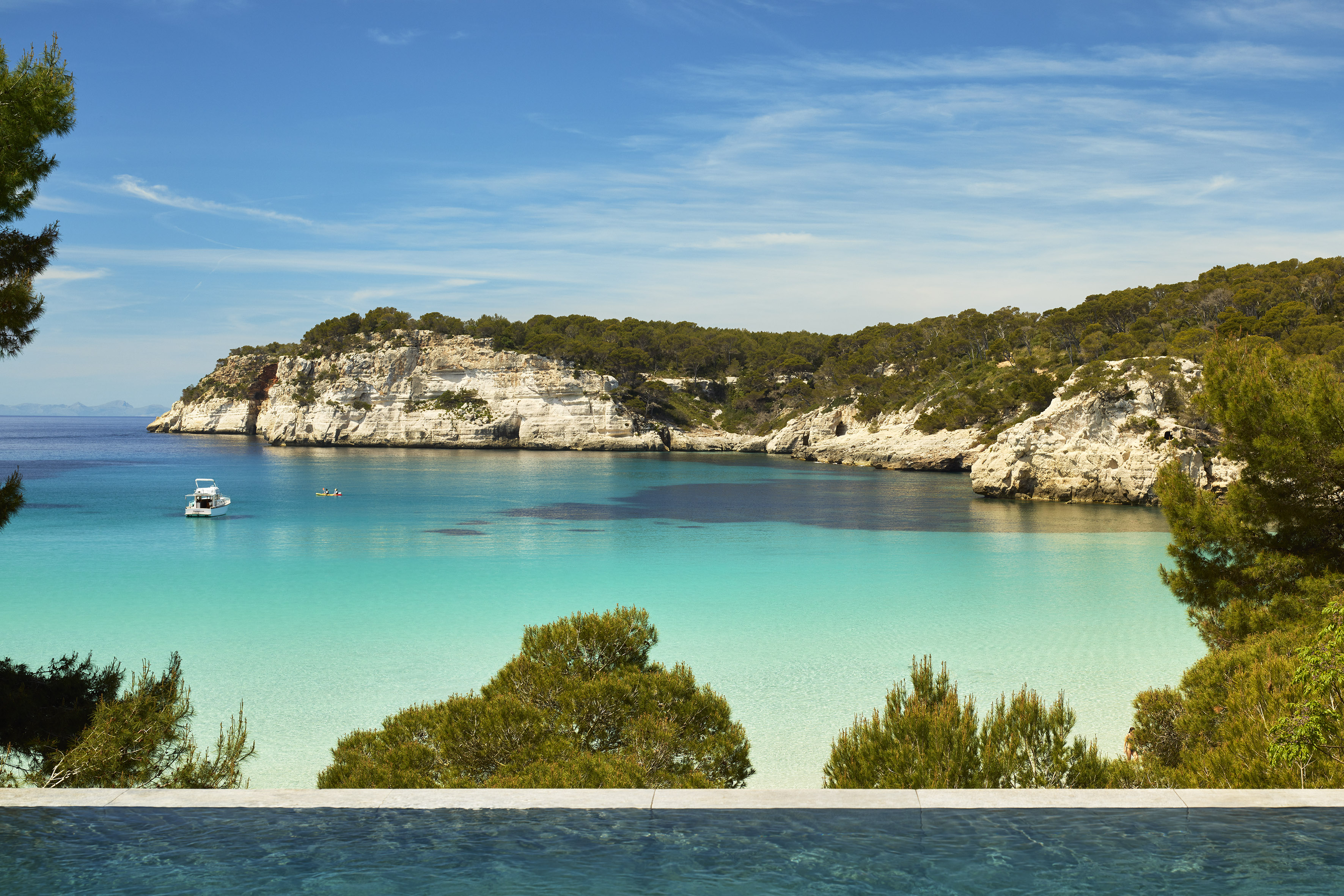 Menorca bay with turquoise water, white cliffs, boat, and kayakers.