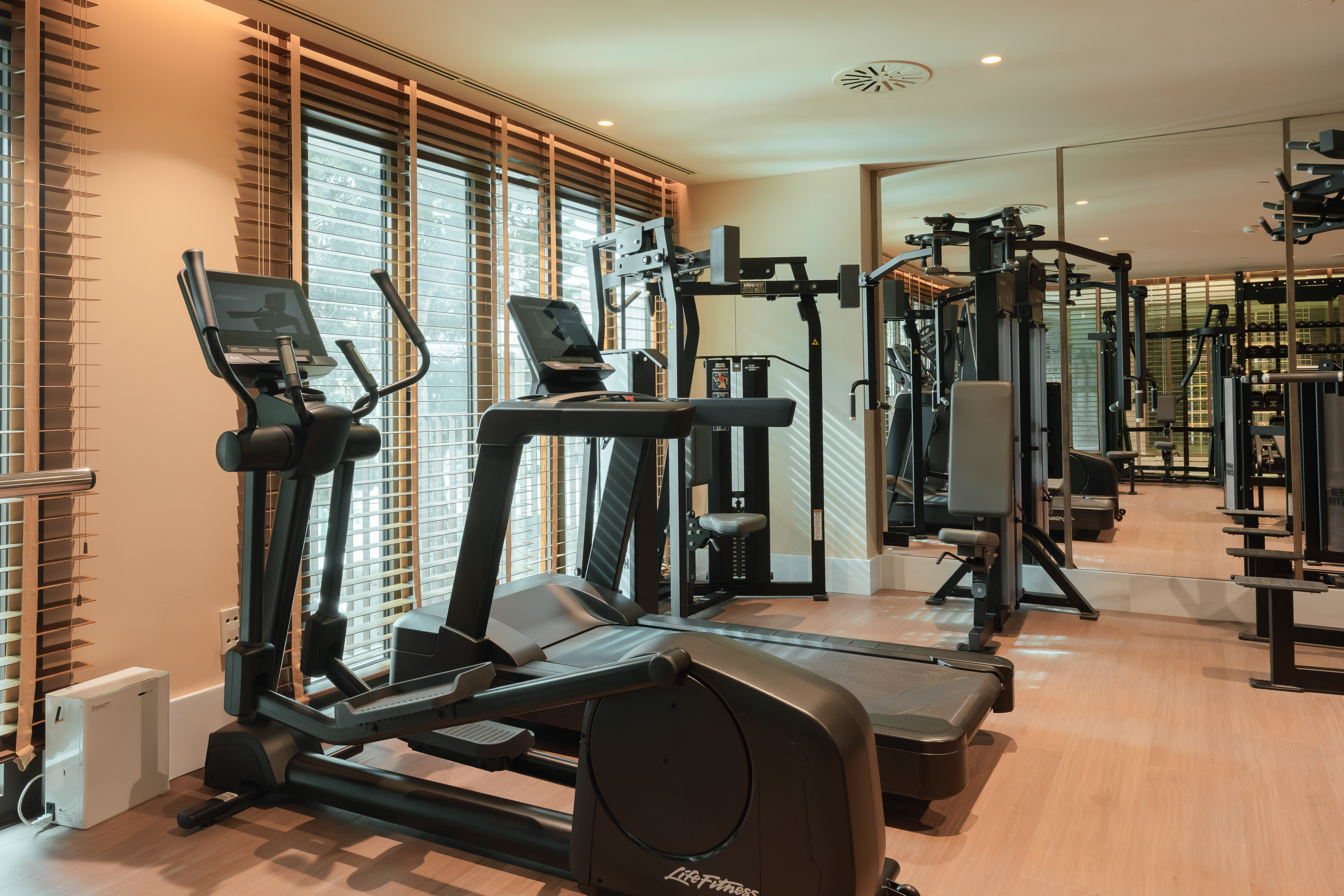 Fitness room with treadmill, elliptical, weight equipment, and window blinds.
