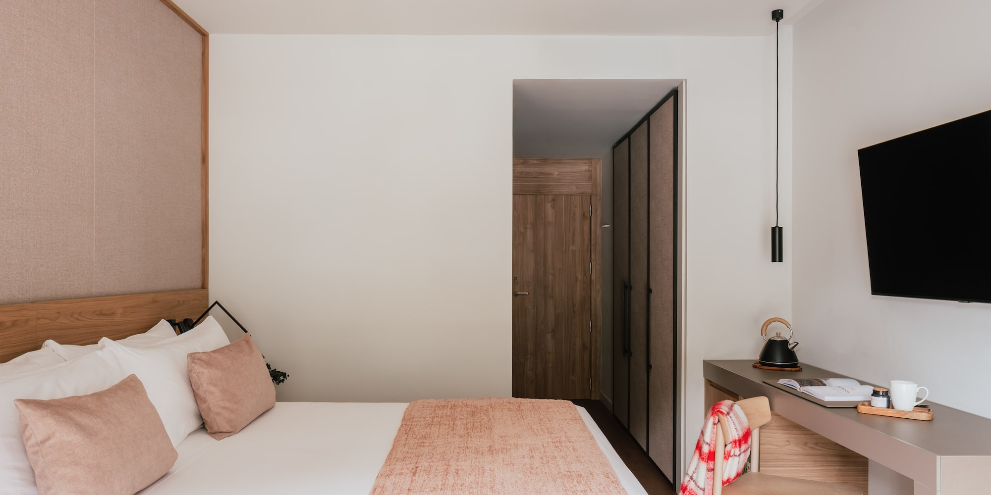Modern hotel bedroom with cozy bed, wood headboard, desk, and flat-screen TV.