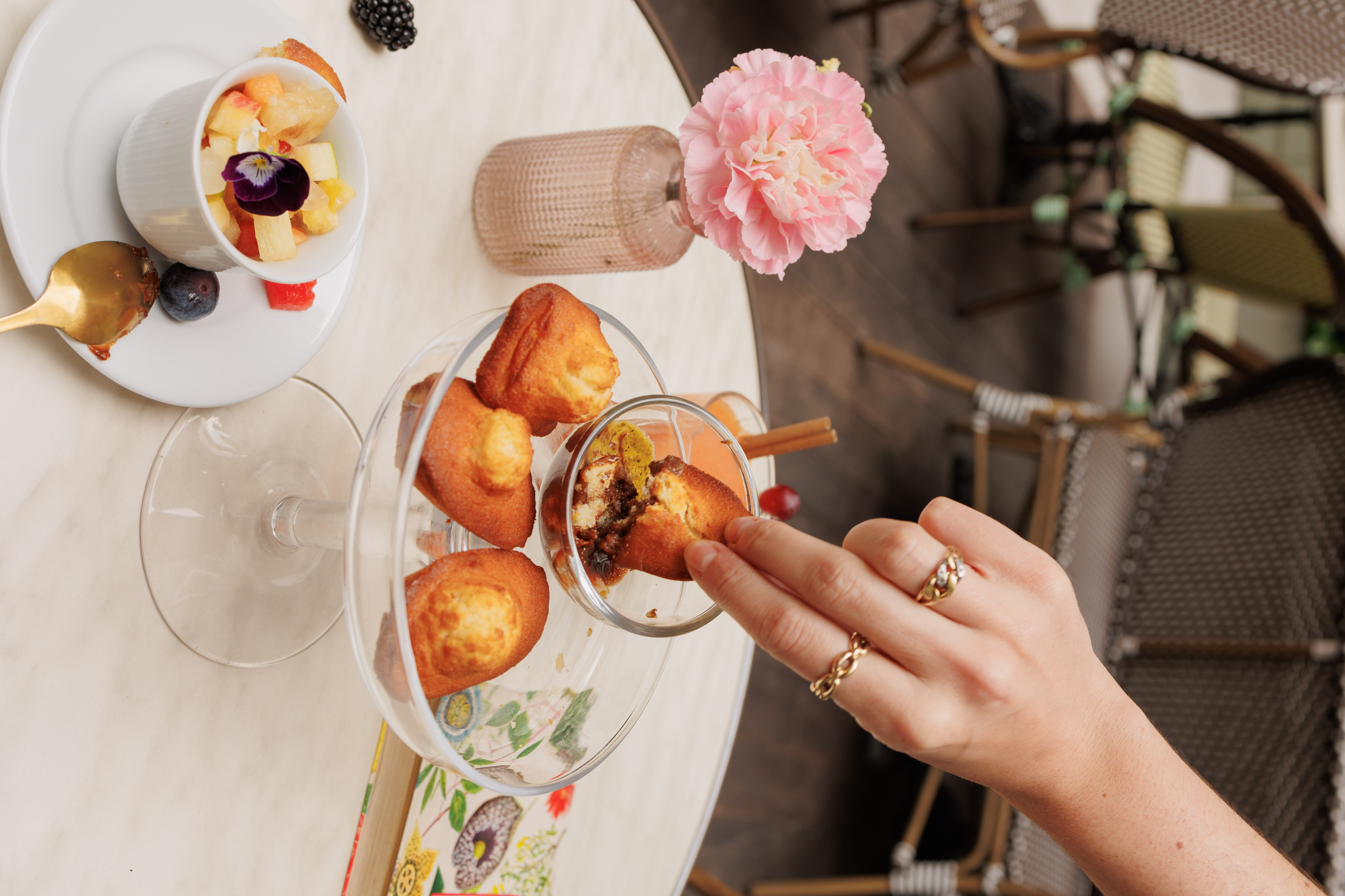 Hand dipping madeleine dessert into sauce, with fruit salad and pink flower on cafe table.