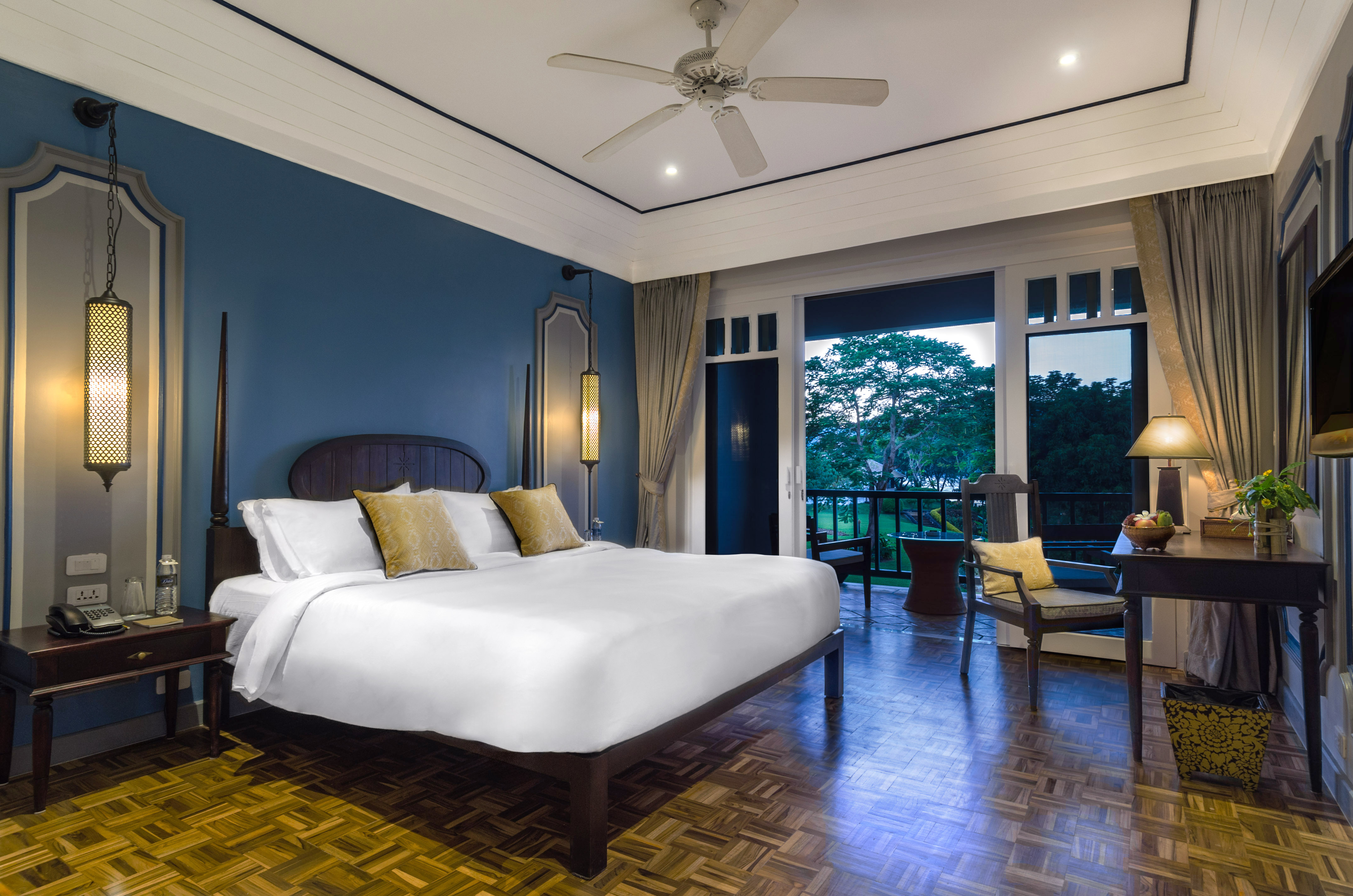 Luxury hotel bedroom with blue walls, king bed, and balcony overlooking lush greenery.