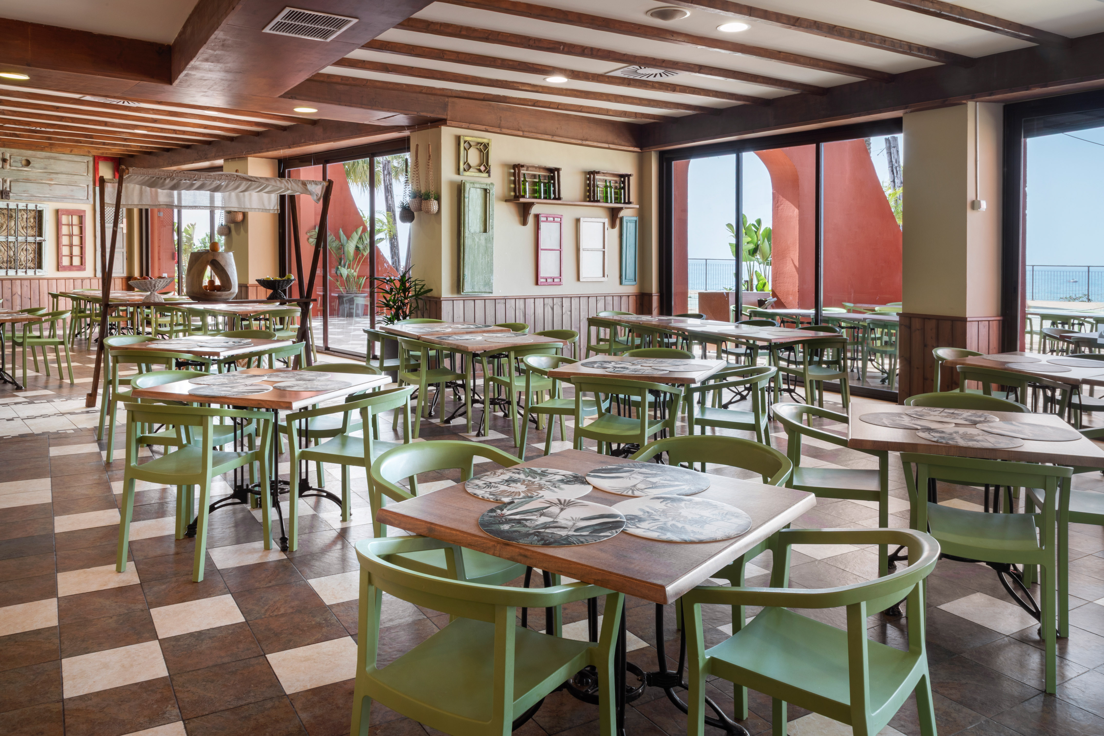 Bright restaurant dining area with green chairs, patterned floor, and sea view.