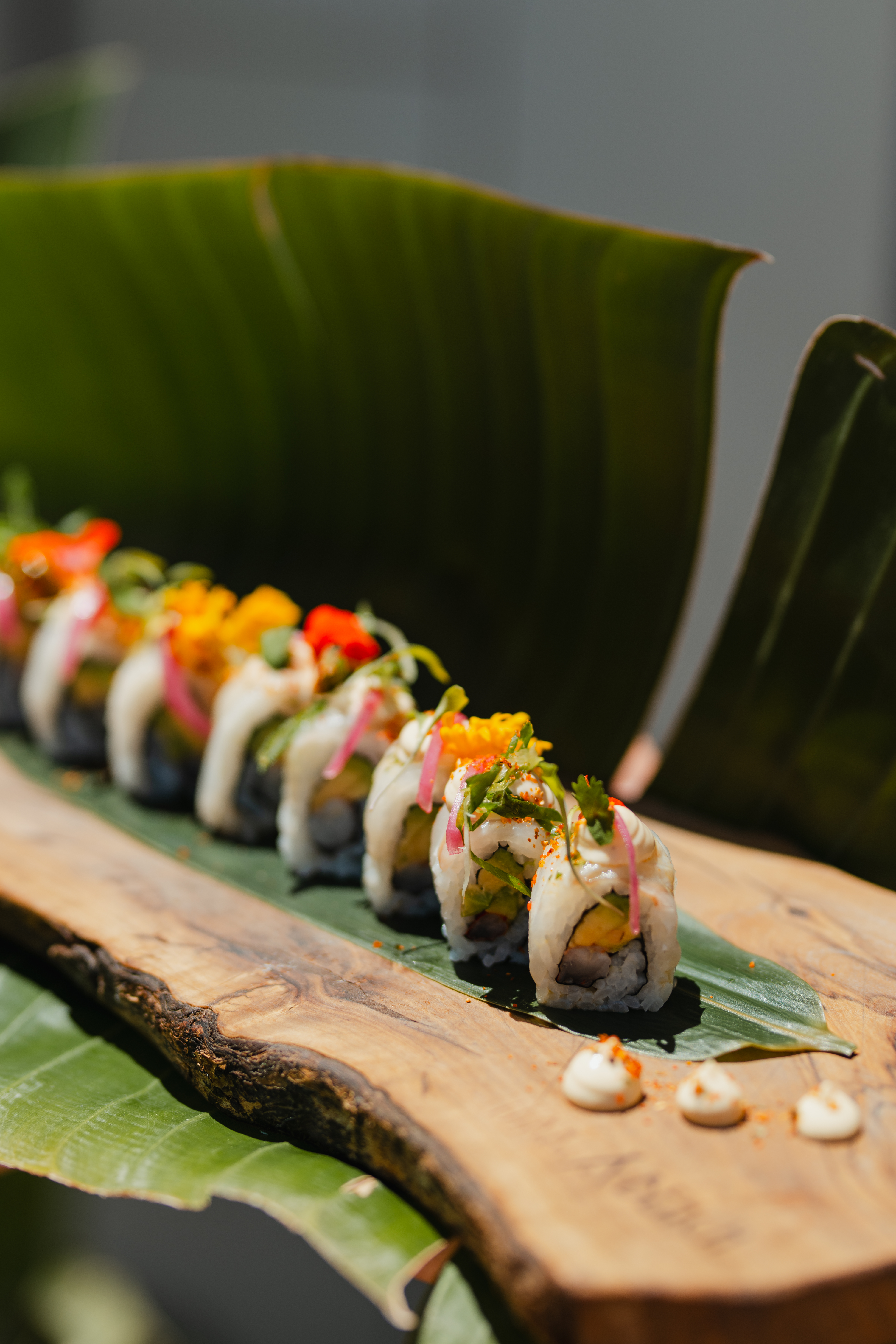 Sushi rolls with colorful toppings on a wooden board and tropical leaves.