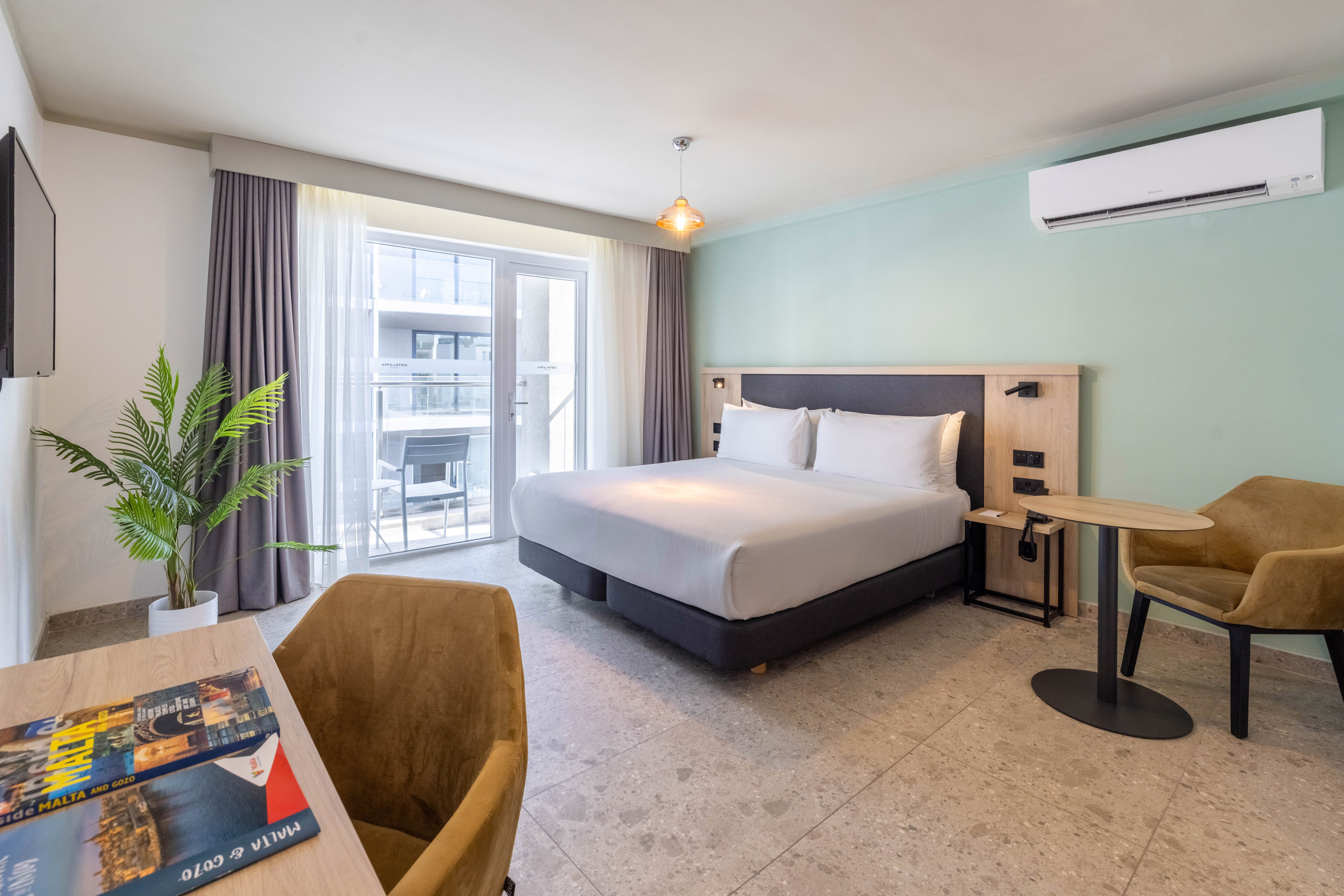 Modern hotel room with king bed, light green wall, balcony access, and desk.