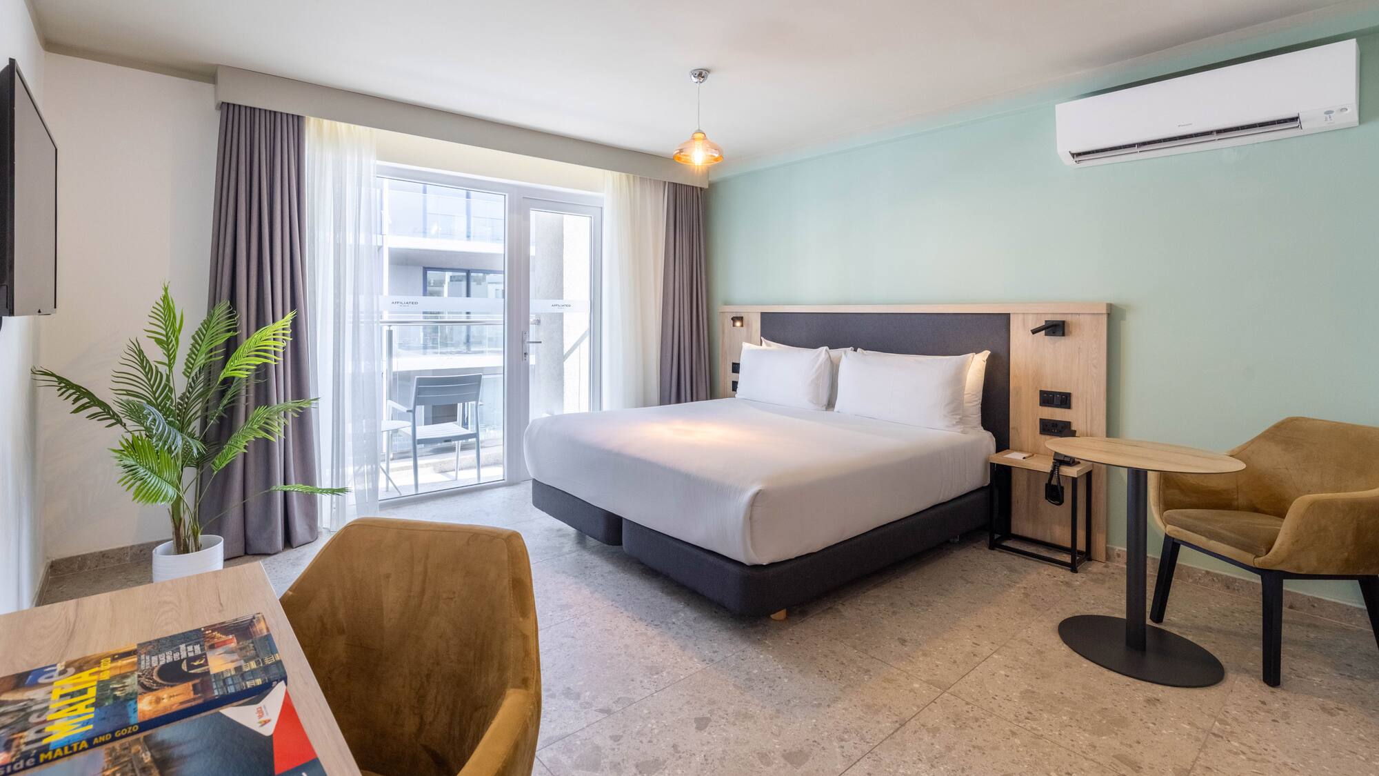 Modern hotel room with king bed, light green wall, balcony access, and desk.