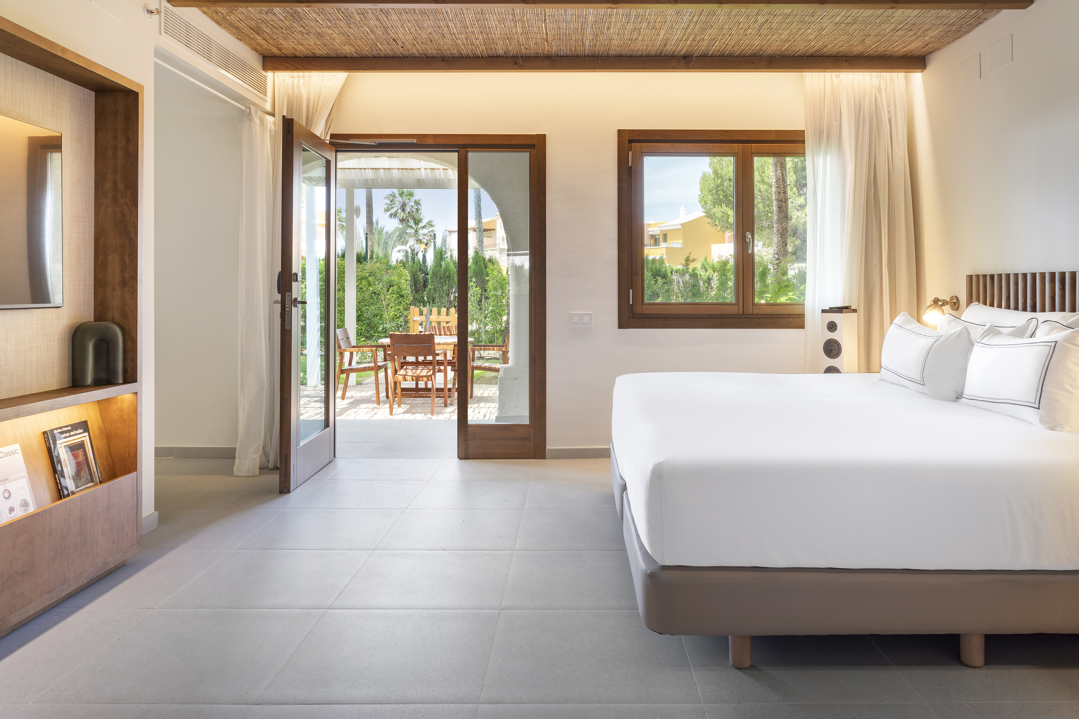 Bright modern bedroom with white bed, patio access, and lush garden view.