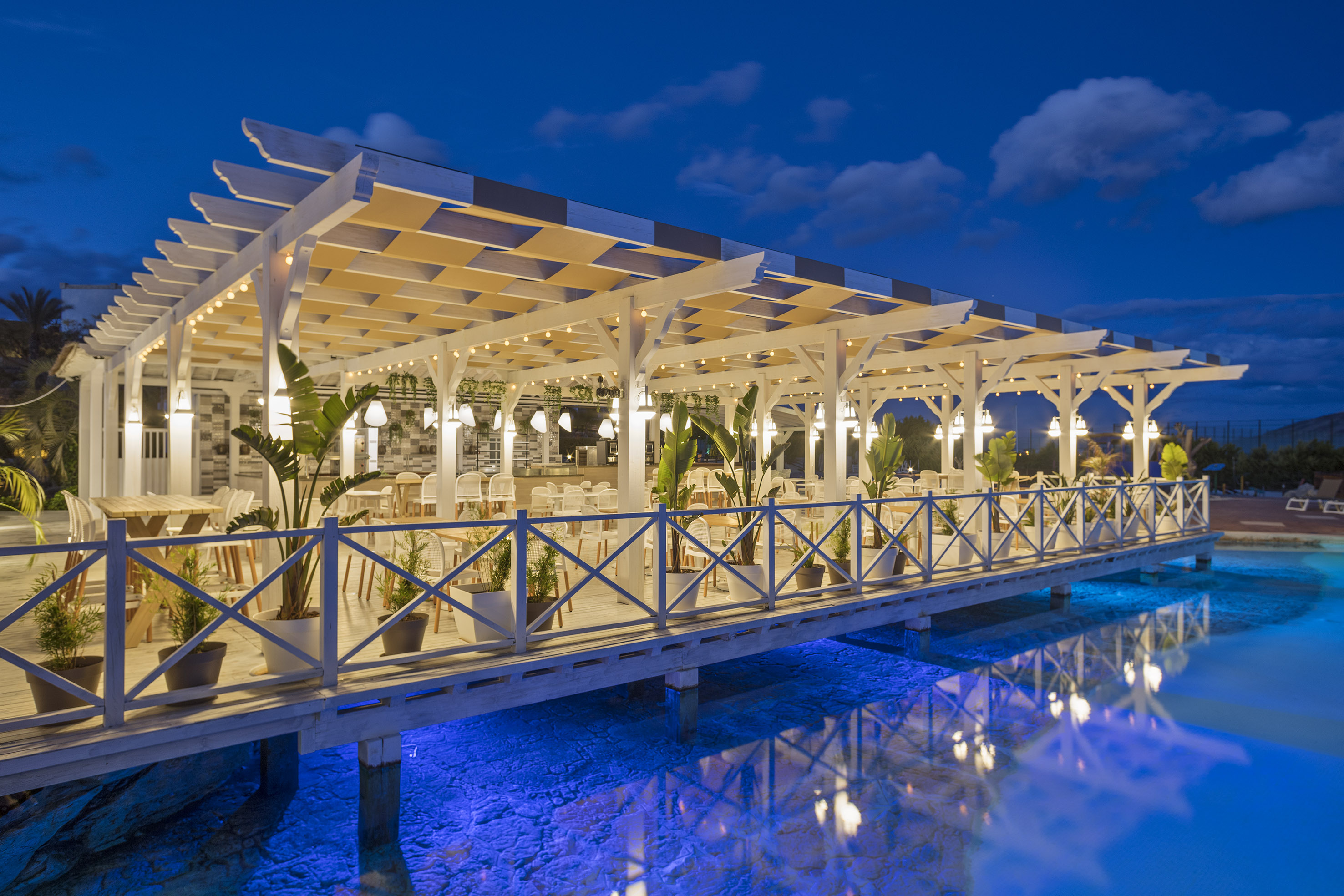 White pergola restaurant over blue water at night, illuminated outdoor dining.