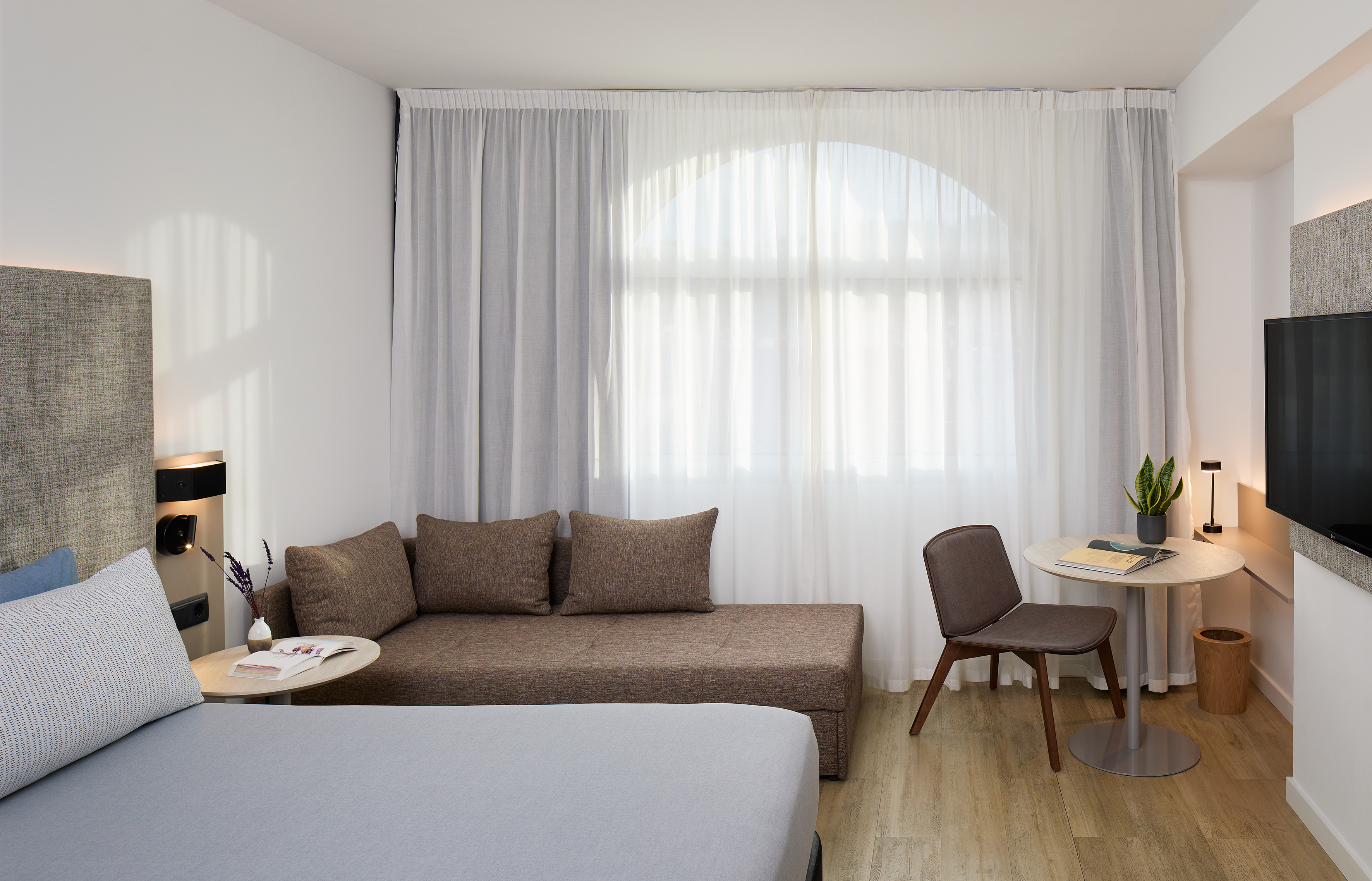 Contemporary hotel room with bed, sofa, tables, and a large arched window.